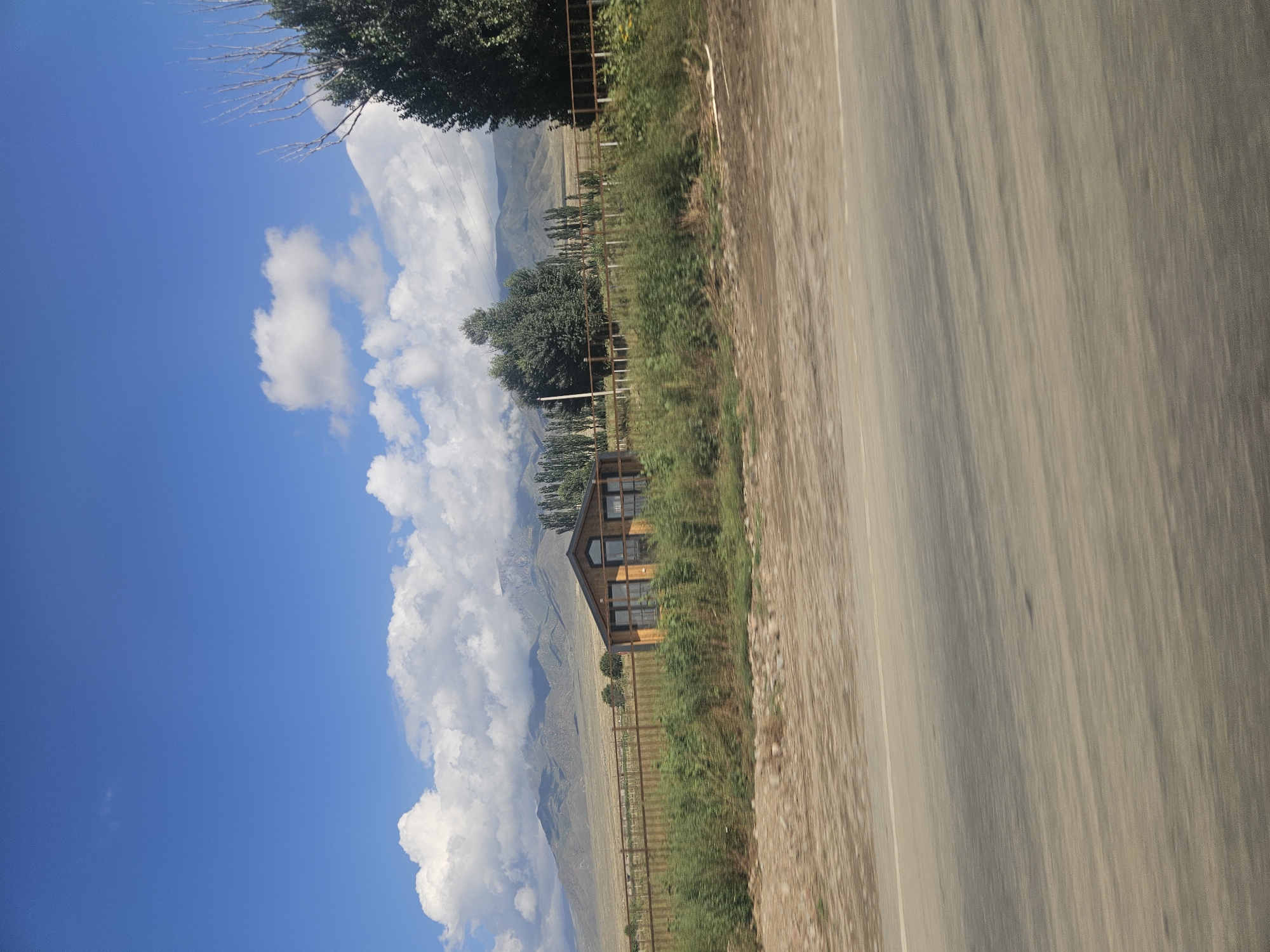 The image depicts a rural landscape in Choktal, Kyrgyzstan, under a bright blue sky with scattered white clouds. In the foreground, a blurred asphalt road stretches across the frame, with a dusty, gravelly shoulder to its left. Beyond the road, a fence runs horizontally, bordering a stretch of green vegetation and a small, wooden house with large windows. In the background, rolling hills and distant mountains are visible, partially obscured by clouds. Several trees, including a tall, bare tree on the right and a cluster of slender trees in the middle, dot the landscape. The overall scene conveys a sense of natural beauty and tranquility.