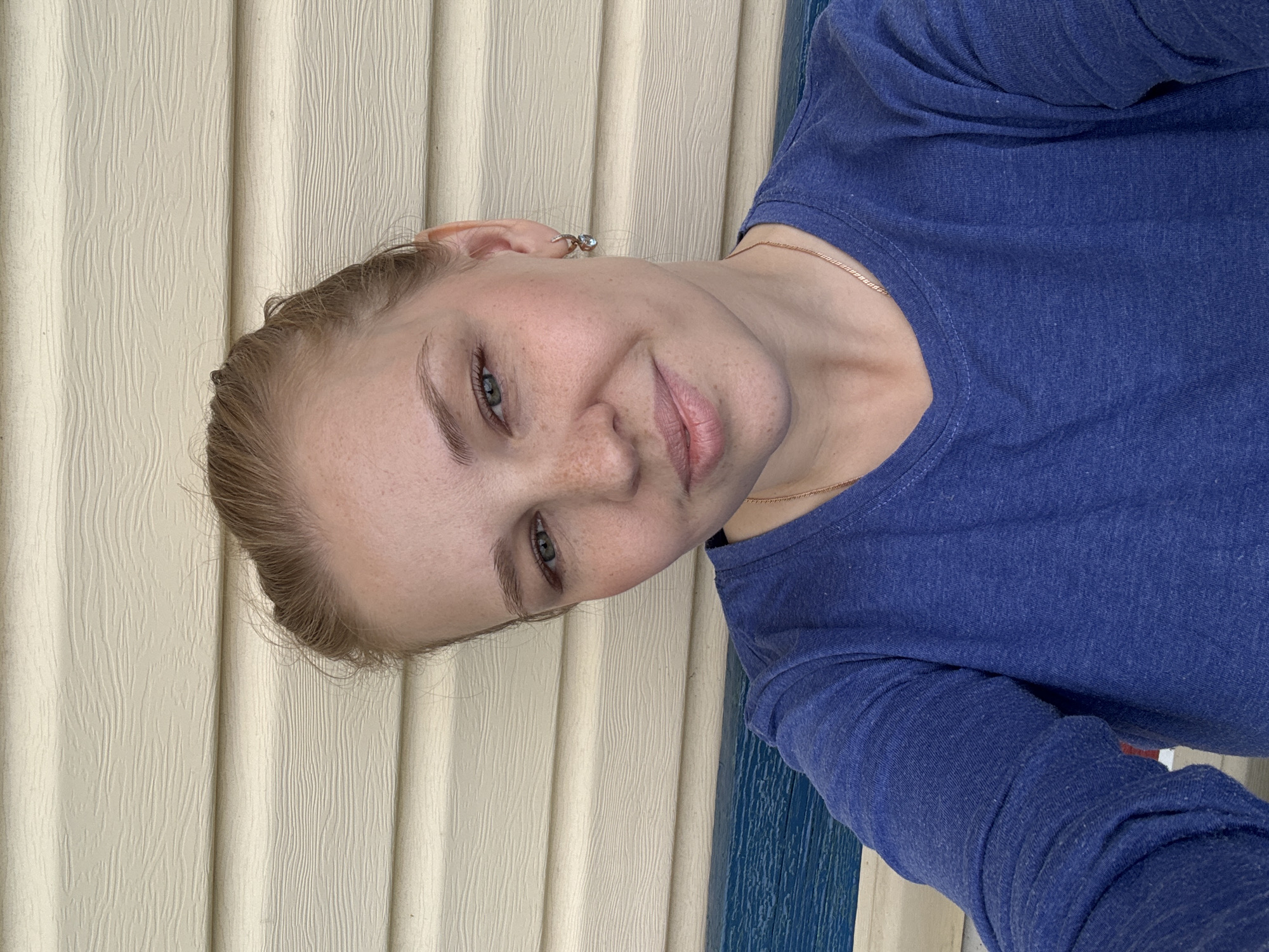 A young woman with blonde hair pulled back and freckles on her face smiles gently at the camera. She is wearing a blue long-sleeved shirt and a delicate necklace. She has a small earring in her left ear. The background consists of horizontal beige siding and a blue railing. This scene appears to be on a porch. The location is identified as Dunilovo, Russia.