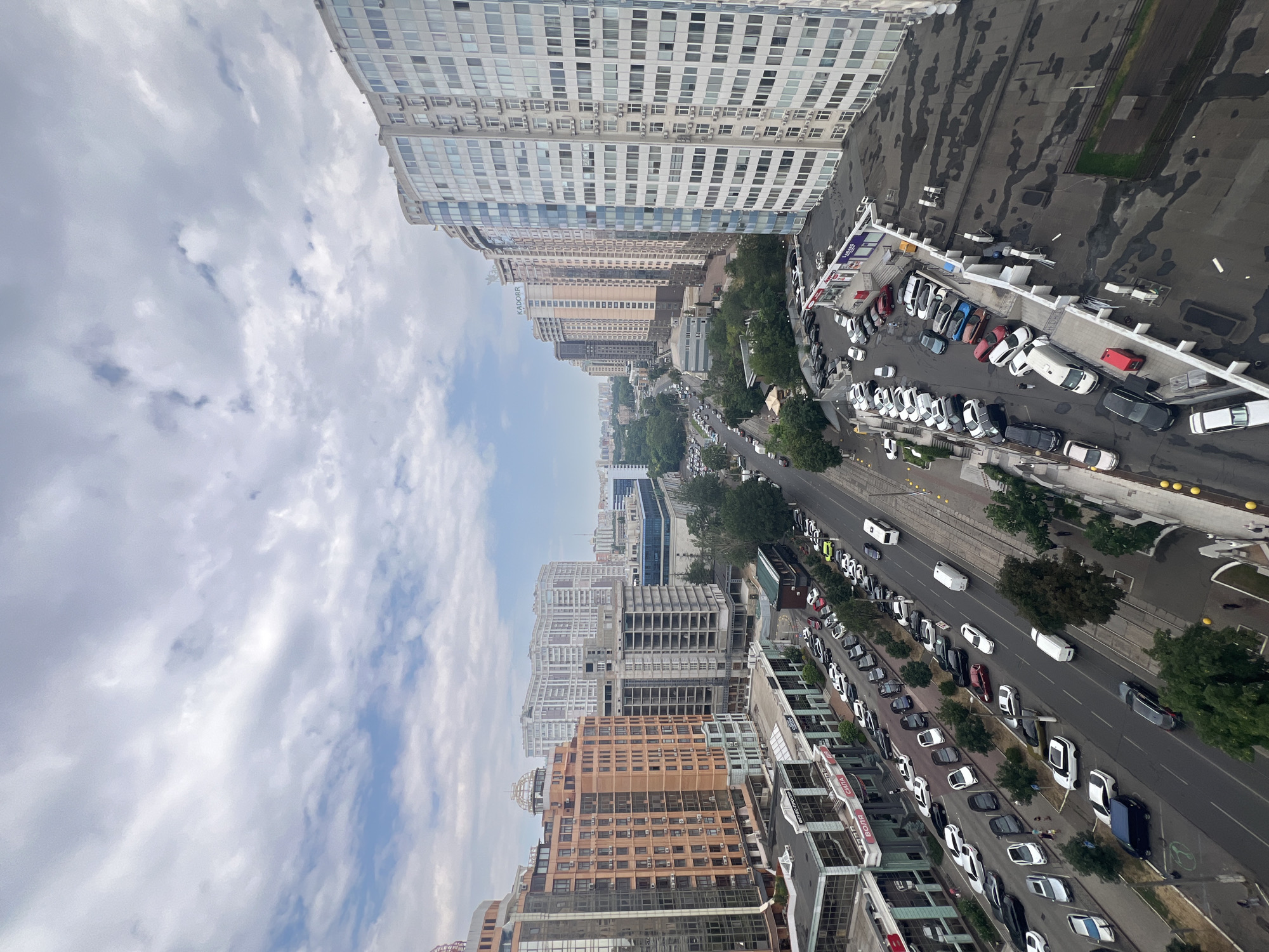 The image captures a panoramic view of a city street in Odesa, Ukraine, on an overcast day. The perspective is from a high vantage point, looking down on a multi-lane road and the surrounding buildings. On the left side of the street, several modern, multi-story buildings are visible. The closest one is a light orange building with many windows and visible air conditioning units. Next to it is a taller, unfinished grey concrete building. Further down the street, a large, white, multi-story residential building dominates the skyline, featuring numerous windows and balconies.
The street itself is busy with traffic. Cars of various colors and models are seen driving and parked along the sides. There are also some larger vehicles, including a white bus or shuttle. Pedestrians are sparsely visible, appearing as small figures on sidewalks and near buildings. The road has multiple lanes, and parking areas are present along the edges of the buildings.
On the right side of the street, more multi-story buildings line the road, including a prominent, tall, curved white building with a blue-tinted glass facade. The rooftops of some lower buildings are also visible, some appearing to be flat with various structures on them. The environment suggests a developed urban area with a mix of residential and commercial structures. The sky is filled with clouds, with patches of blue sky peeking through, indicating a partly cloudy or overcast weather condition.