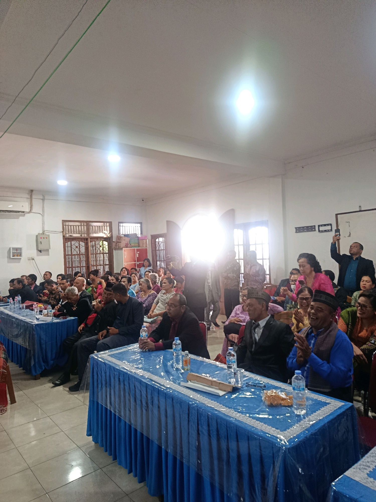 The image depicts an indoor gathering of people in Babelan, Indonesia. The scene appears to be a formal event or ceremony taking place in a room with tiled floors and white walls. A bright overhead light illuminates the space, casting reflections on the floor.
In the foreground, two tables draped with blue tablecloths and transparent plastic covers are set up. Various items are placed on the tables, including bottles of water, glasses, and what appears to be snacks or offerings. People are seated at these tables and also in the background, suggesting an audience or attendees.
The individuals in the image are dressed in a mix of formal and traditional attire. Many men are wearing suits, some with traditional caps, while women are in dresses or blouses and skirts, some adorned with headscarves or patterned fabrics. Some individuals are actively engaged, such as a man in the background holding up a phone, possibly recording or taking pictures. Others are seated and observing the event. The overall atmosphere suggests a community event, possibly a religious service, a celebration, or a formal meeting. The presence of windows with wooden frames and decorative glass indicates the architectural style of the building.