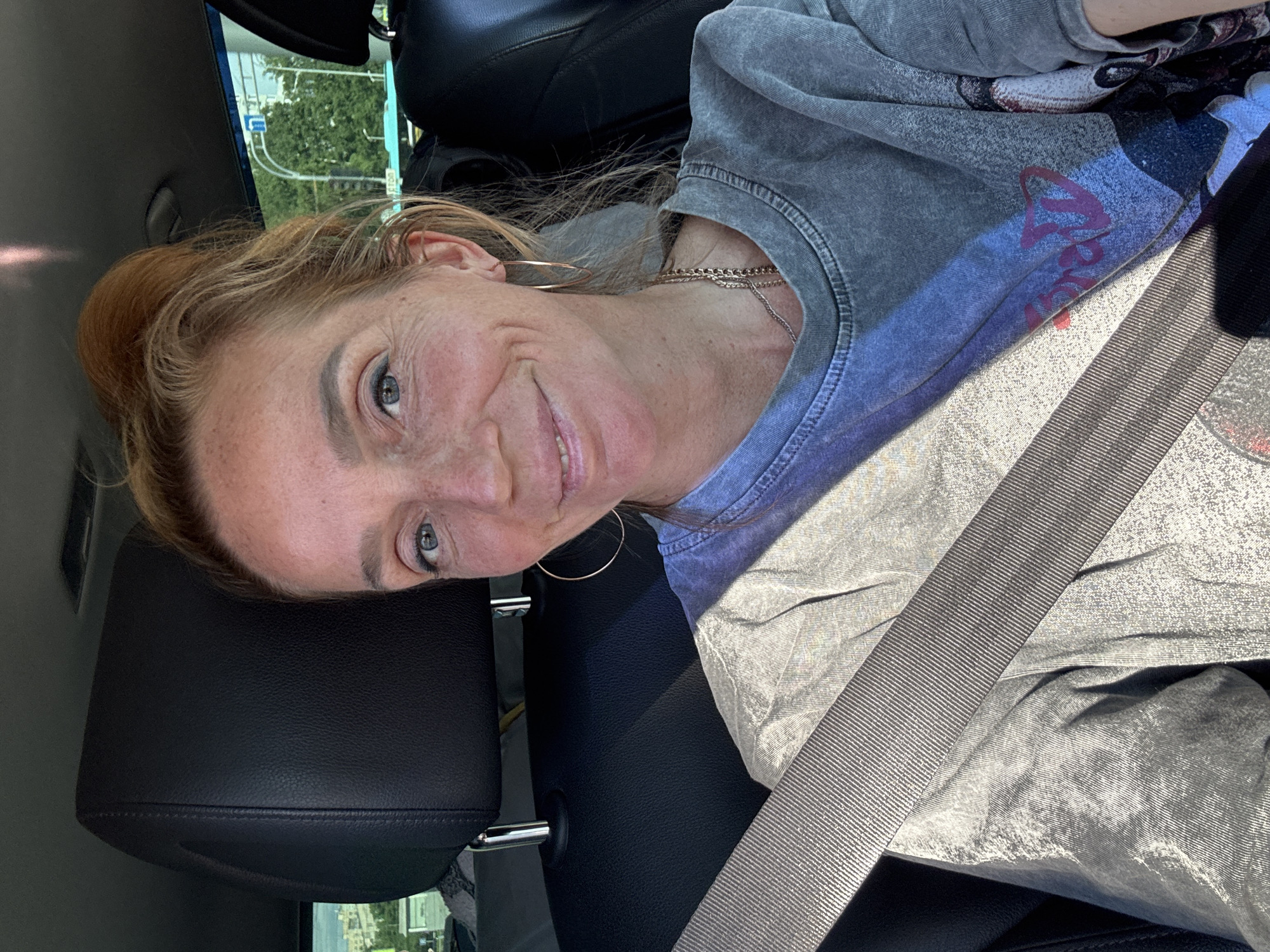 An adult female with reddish-brown hair tied in a bun, fair skin, and prominent freckles, smiles directly at the camera. She wears a grey, stone-washed short-sleeved t-shirt with a partial graphic print featuring a cartoon character's eye and pink lettering. Gold-toned hoop earrings and multiple thin gold-toned necklaces are visible. She is seated in the front passenger seat of a vehicle, with a dark grey seatbelt buckled across her chest. The car's interior consists of black leather upholstery visible on the headrest and seat. Through the windows, an outdoor urban environment is visible, featuring buildings, green trees, and a road sign or traffic structure under bright daylight. The scene takes place in Yukki, Russia.