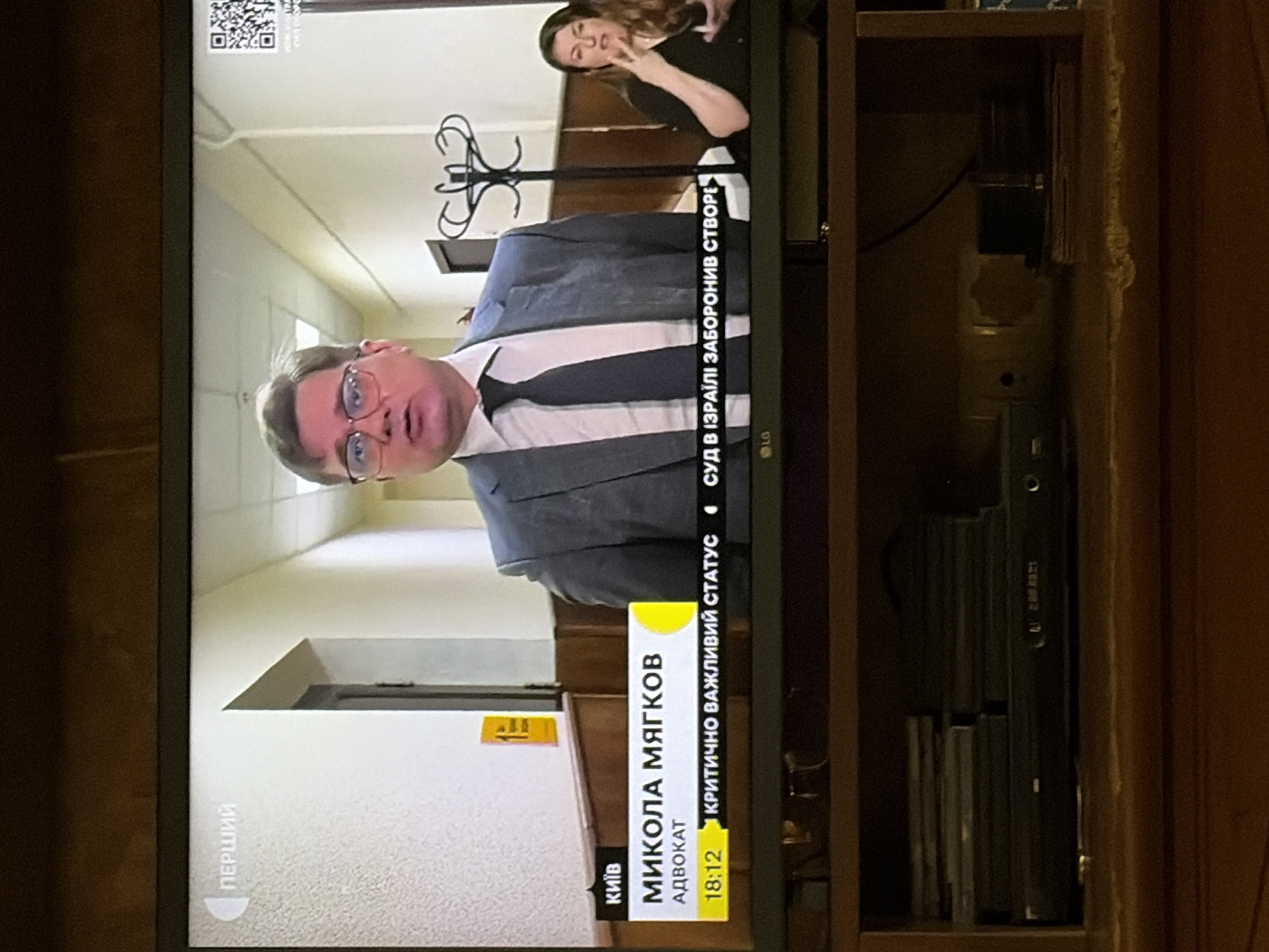 The image shows a television screen displaying a news broadcast. On the screen, a man wearing glasses and a suit is speaking. Below him, text indicates his name is "МИКОЛА МЯГКОВ" and his profession is "АДВОКАТ" (lawyer). The broadcast originates from "КИЇВ" (Kyiv). There is also a QR code on the top right of the screen. To the right of the main speaker, a woman is visible, seemingly an interpreter or commentator, using sign language. The lower part of the image shows a shelf with several DVDs or cases and a black electronic device, possibly a DVD player. The overall setting appears to be indoors, with a hallway visible behind the speakers on the television.