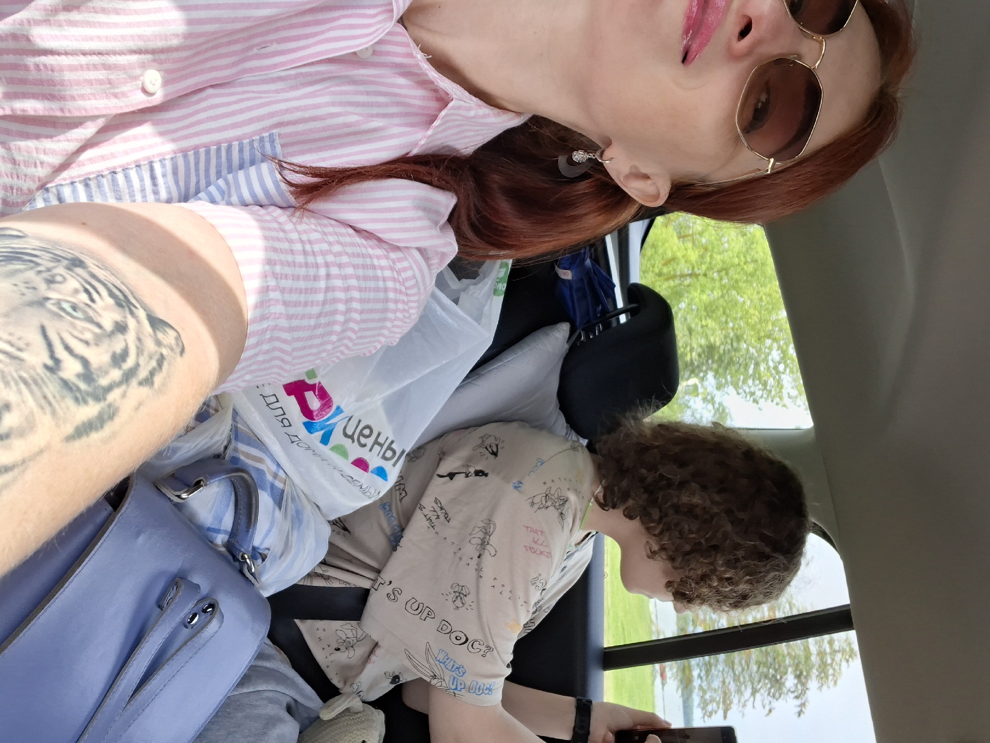 A woman with auburn hair and sunglasses is in the front passenger seat of a car, looking at the camera. She is wearing a pink and white striped shirt and a tattoo of a tiger's face is visible on her arm. A younger person with curly brown hair is in the back seat, holding a phone. The car is filled with bags and luggage, suggesting travel. The background outside the window shows trees and a body of water, possibly Lake Myadzyel in Myadzyel, Belarus. The lighting indicates it is daytime, with sunlight filtering into the car.