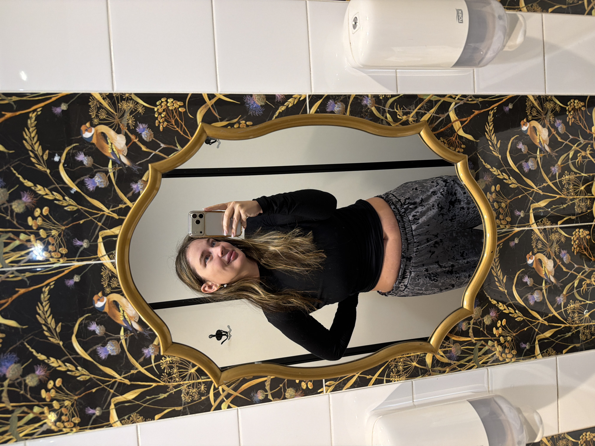 An image, rotated 90 degrees counter-clockwise, depicts a woman taking a mirror selfie in what appears to be a stylish restroom in Moscow, Russia.
The central subject is the woman, reflected in an ornate, gold-framed mirror. She has long, dark hair with lighter streaks and is wearing a black long-sleeved crop top and patterned grey pants. She holds a light-colored smartphone to capture her reflection, smiling playfully with her tongue subtly sticking out.
The restroom environment features a distinctive wall design. Behind the mirror, the wall is covered with dark wallpaper or tiles adorned with a repeating pattern of golden foliage, purple thistles, and small birds resembling European goldfinches. Above and below this decorative section, the walls are covered with clean white square tiles. Two white and grey dispensers, likely for soap or paper towels, are mounted on the tiled wall; one prominently displays the brand name "TORK." A black wall hook is also visible in the mirror's reflection behind the woman.
The overall scene suggests a well-decorated, possibly upscale, public or commercial establishment, with the woman expressing a lighthearted and happy mood through her selfie. The time of day is not discernible, as the setting is indoors with artificial lighting.