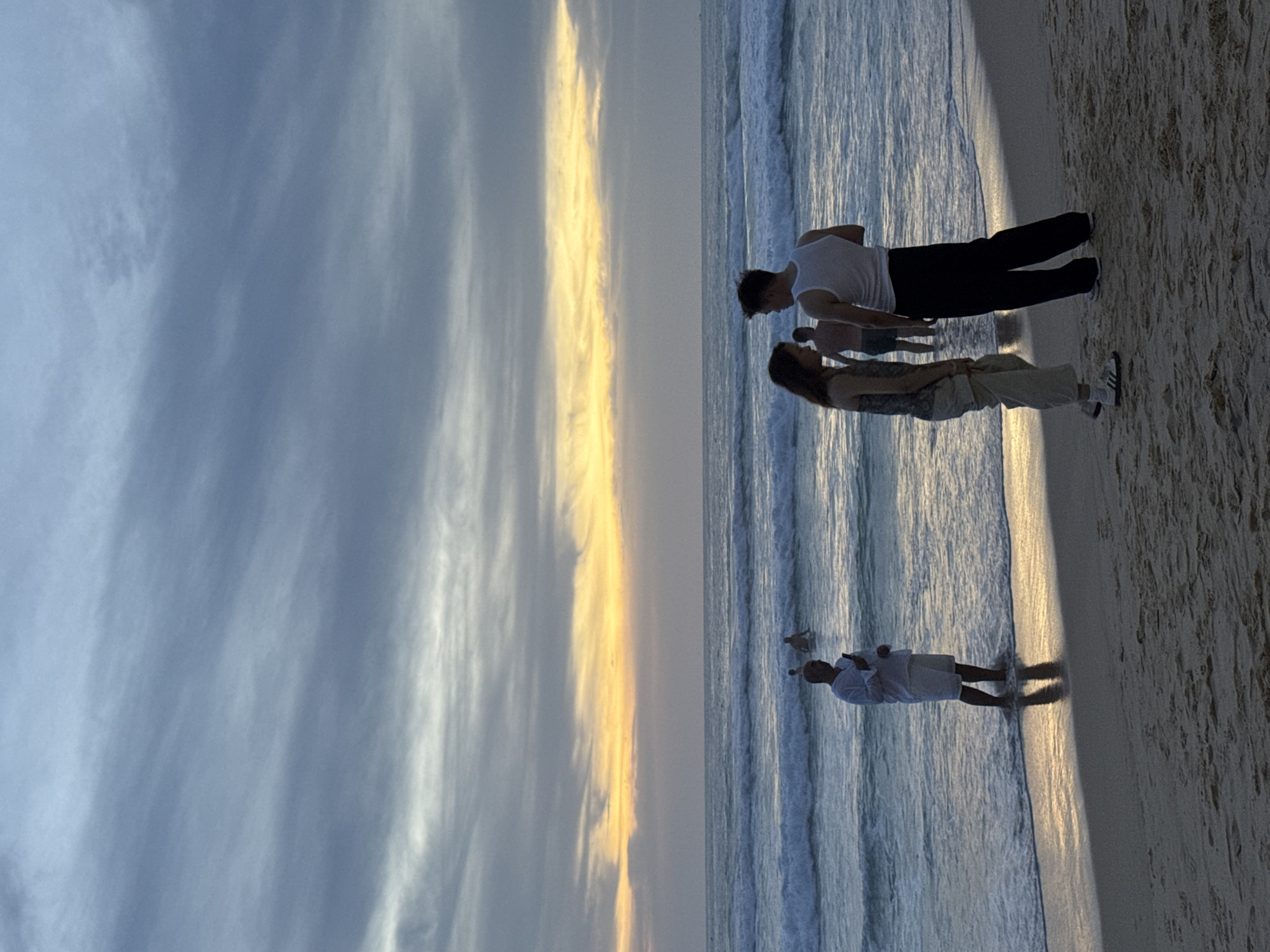 At Karon Beach, Thailand, during twilight, a man stands in the shallow ocean, facing away from the camera and holding a phone. In the background, several individuals are wading in the water. Closer to the shore, a couple is engaged in a conversation on the sandy beach. The sky is filled with streaks of gray and white clouds, with a pale yellow glow emanating from the horizon, suggesting the sun has recently set. The waves are moderate and break onto the shore, creating a foamy white surf. The sand is a light brown color, and the lighting is dim, characteristic of dusk.