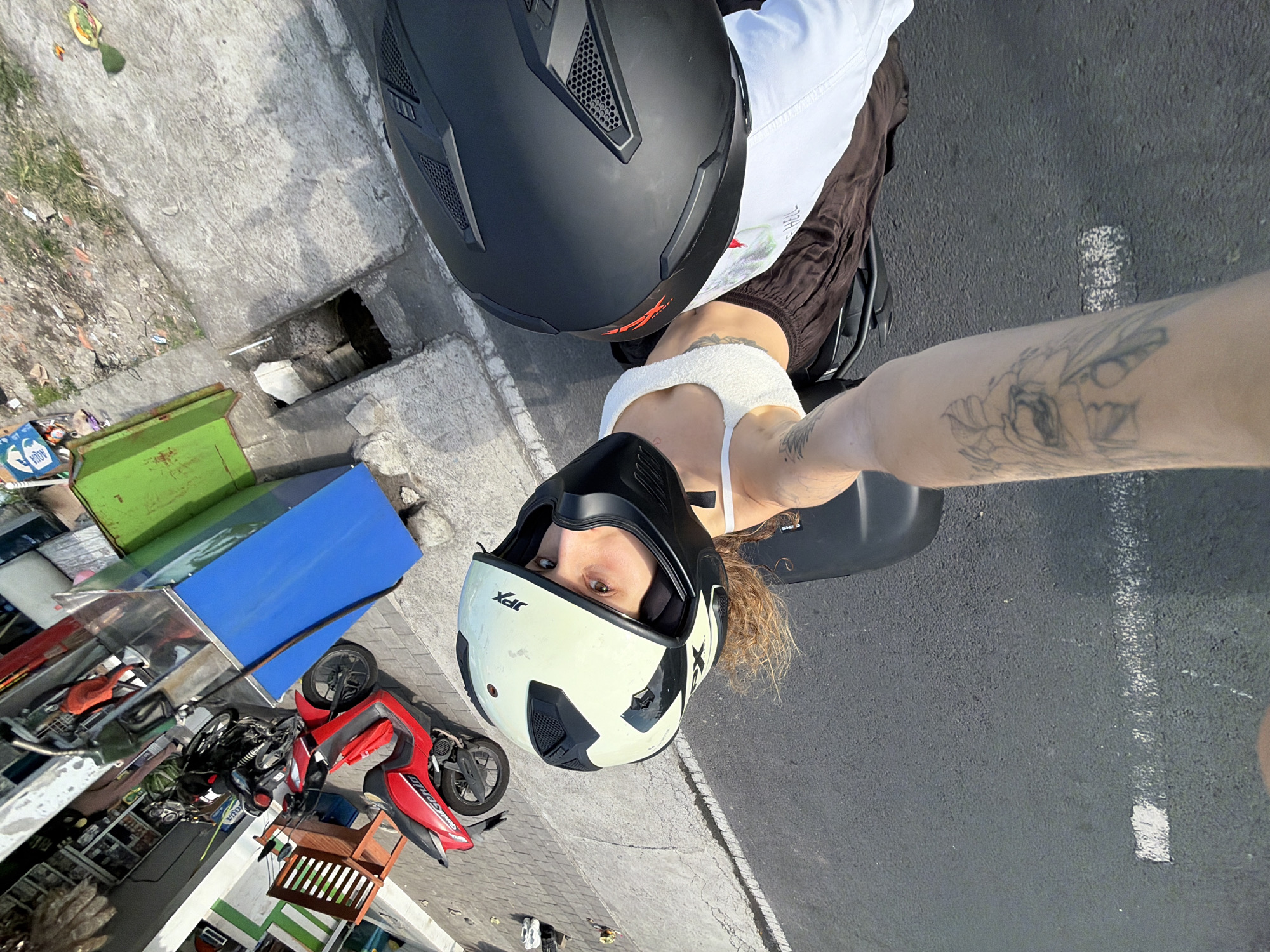 A selfie taken from the perspective of a passenger on a motorbike. The passenger, wearing a white tank top and brown shorts, has a black helmet on their head, and their arm, adorned with tattoos, is extended out. The driver, wearing a light-colored helmet, is partially visible. The motorbike is red and parked on a paved road in North Kuta, Indonesia. The background shows a street with a green dumpster and a blue gas pump.