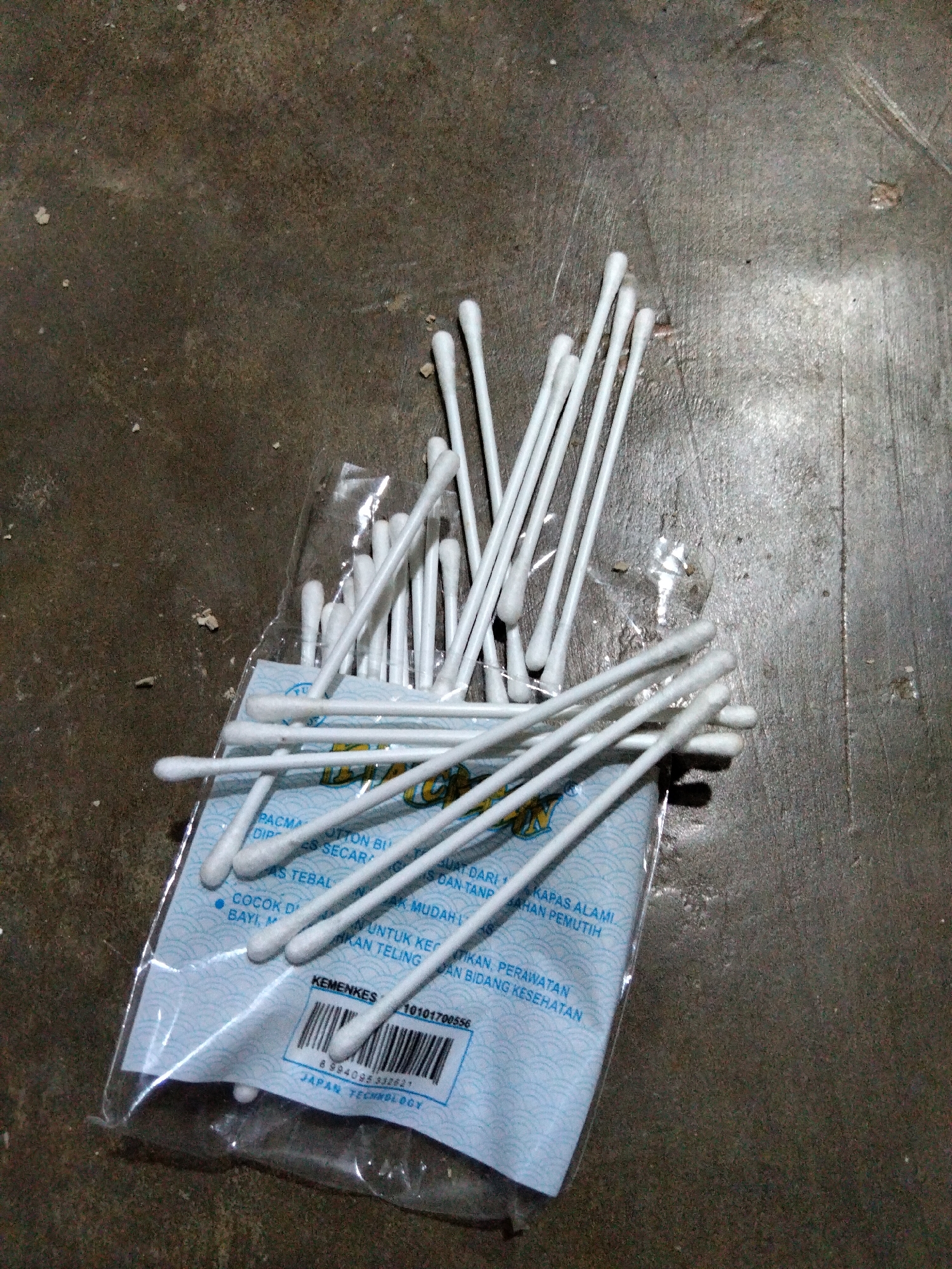 This media file shows a pile of cotton swabs in plastic packaging, lying on a textured, dark surface that appears to be concrete or asphalt. The cotton swabs are white with light brown tips and are scattered across the packaging, with some extending out from it. The packaging itself is light blue with a wave-like pattern and some text in a foreign language. A barcode is visible on the packaging, along with the text "KEMENKES" and "JAPAN TECHNOLOGY." The lighting suggests it is daytime, possibly outdoors, given the rough surface and scattered debris. There are no people or animals visible in the image. The scene is static, with the cotton swabs simply resting on the ground. The overall impression is one of everyday objects in a somewhat utilitarian setting, possibly a storage area or a public space in Benuaraja, Indonesia.