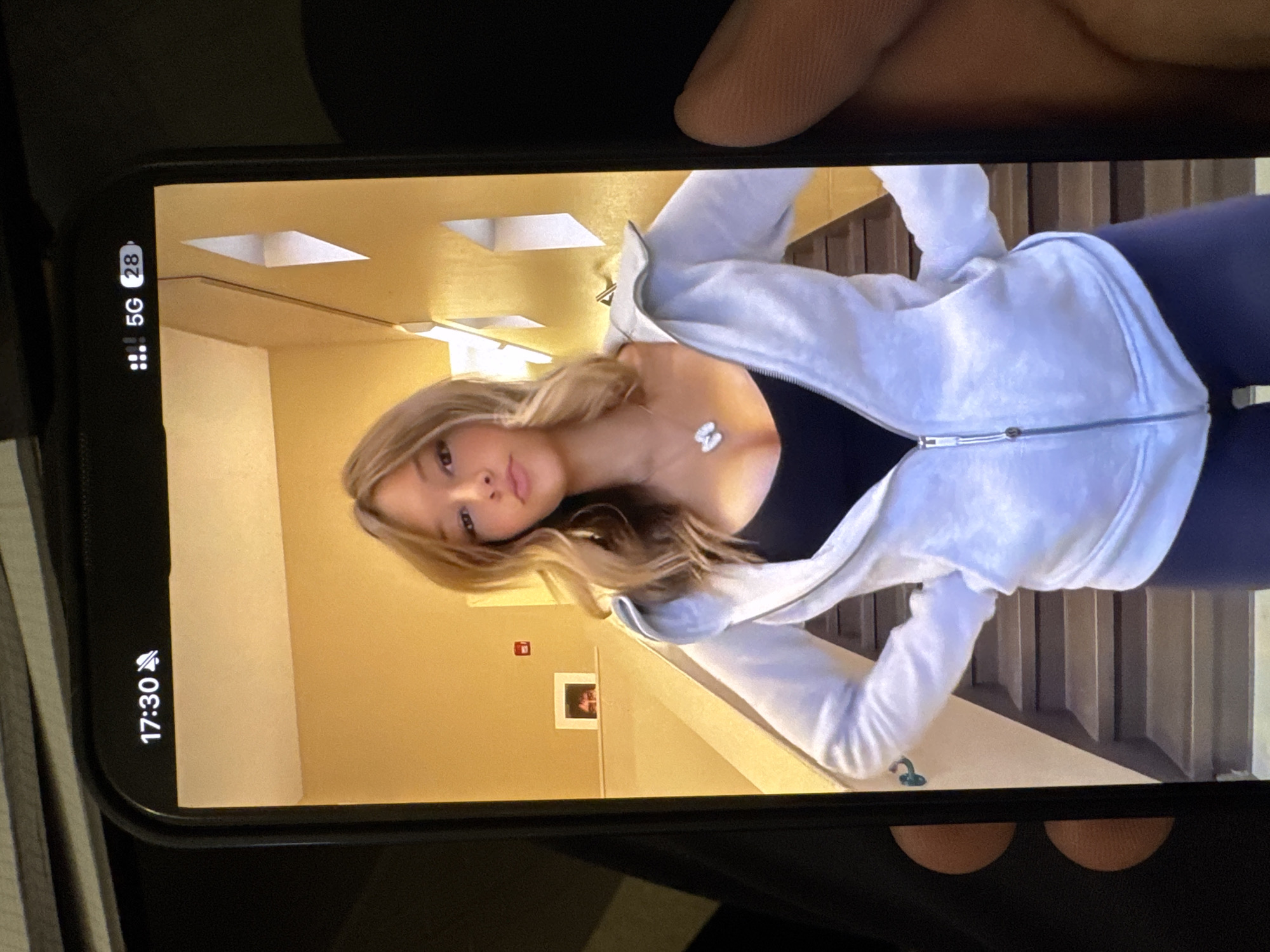 A person is holding a smartphone displaying a photo of a blonde-haired woman with a white zip-up jacket and dark leggings in a hallway. The hallway has yellow walls and geometric light fixtures. A framed picture and a red fire safety sign are visible on the wall behind the woman. The smartphone display shows the time as 17:30 and 5G network connectivity. The location cannot be ascertained as Yerevan, Armenia from the image alone.