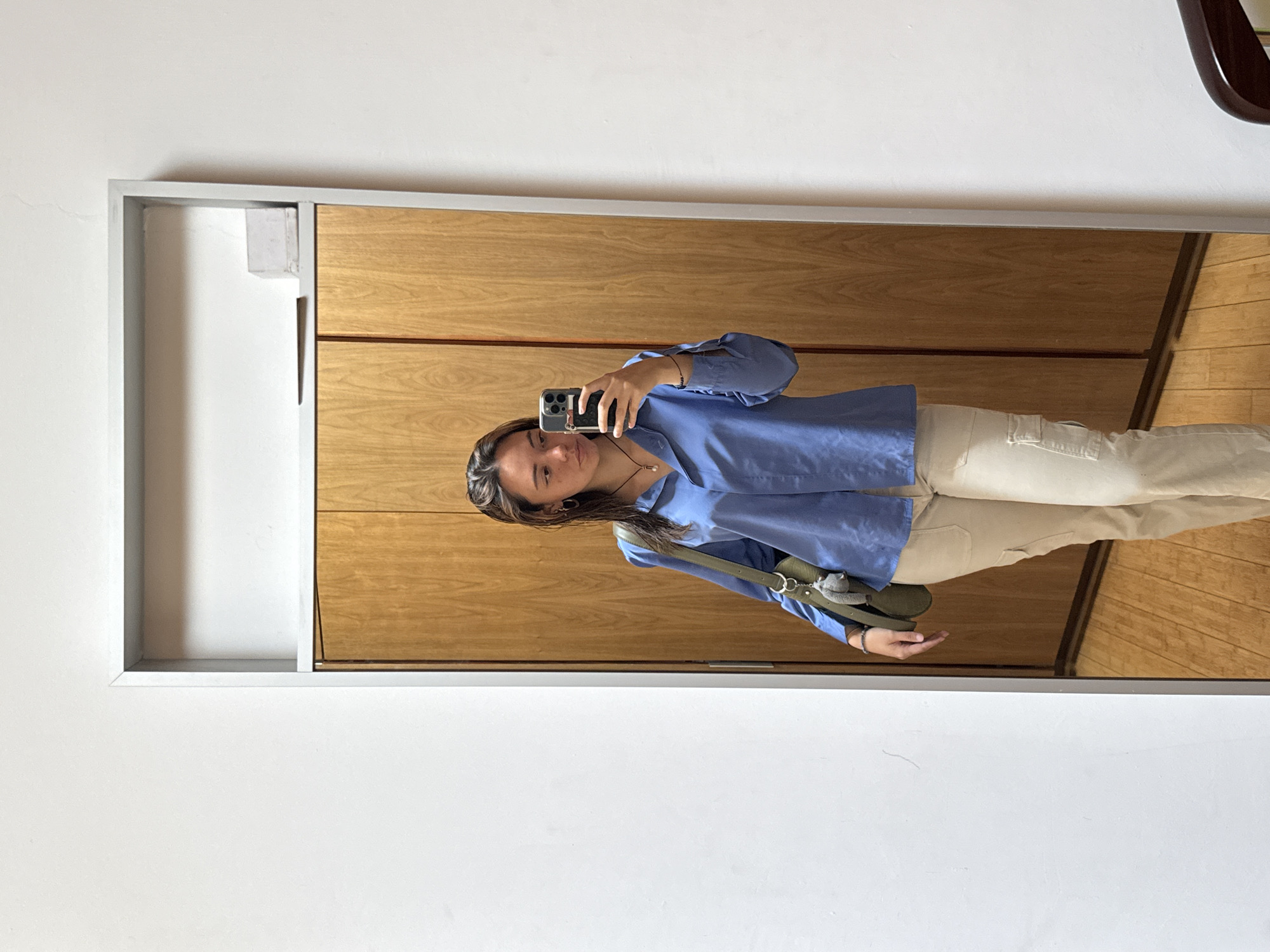A woman is taking a selfie in a mirror. The mirror is framed in silver and is attached to a wall that appears to be white. The wall behind the mirror is paneled with wood. The woman is wearing a blue button-down shirt and light-colored pants. She has a green bag slung over her shoulder. Her hair is brown and she is wearing a necklace. The location appears to be indoors. There is no information available to confirm the location in the city of San Bernardino Tlaxcalancingo, Mexico.