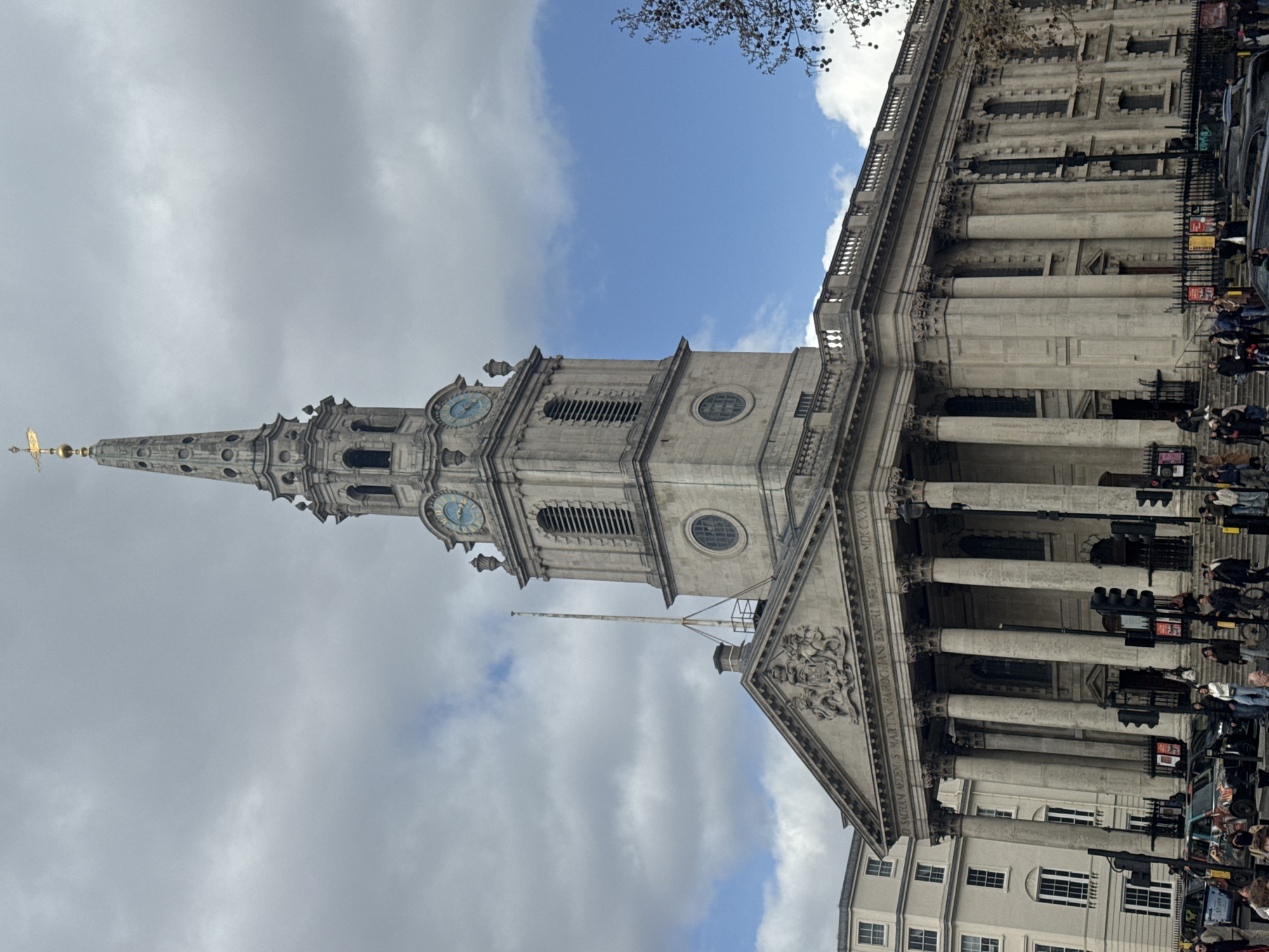 The image shows St. Martin-in-the-Fields church in London, United Kingdom. The church's steeple is visible against a partly cloudy sky. The building features a portico with columns, and people are seen on the sidewalk in front of it. Traffic lights and vehicles are also visible near the building.
