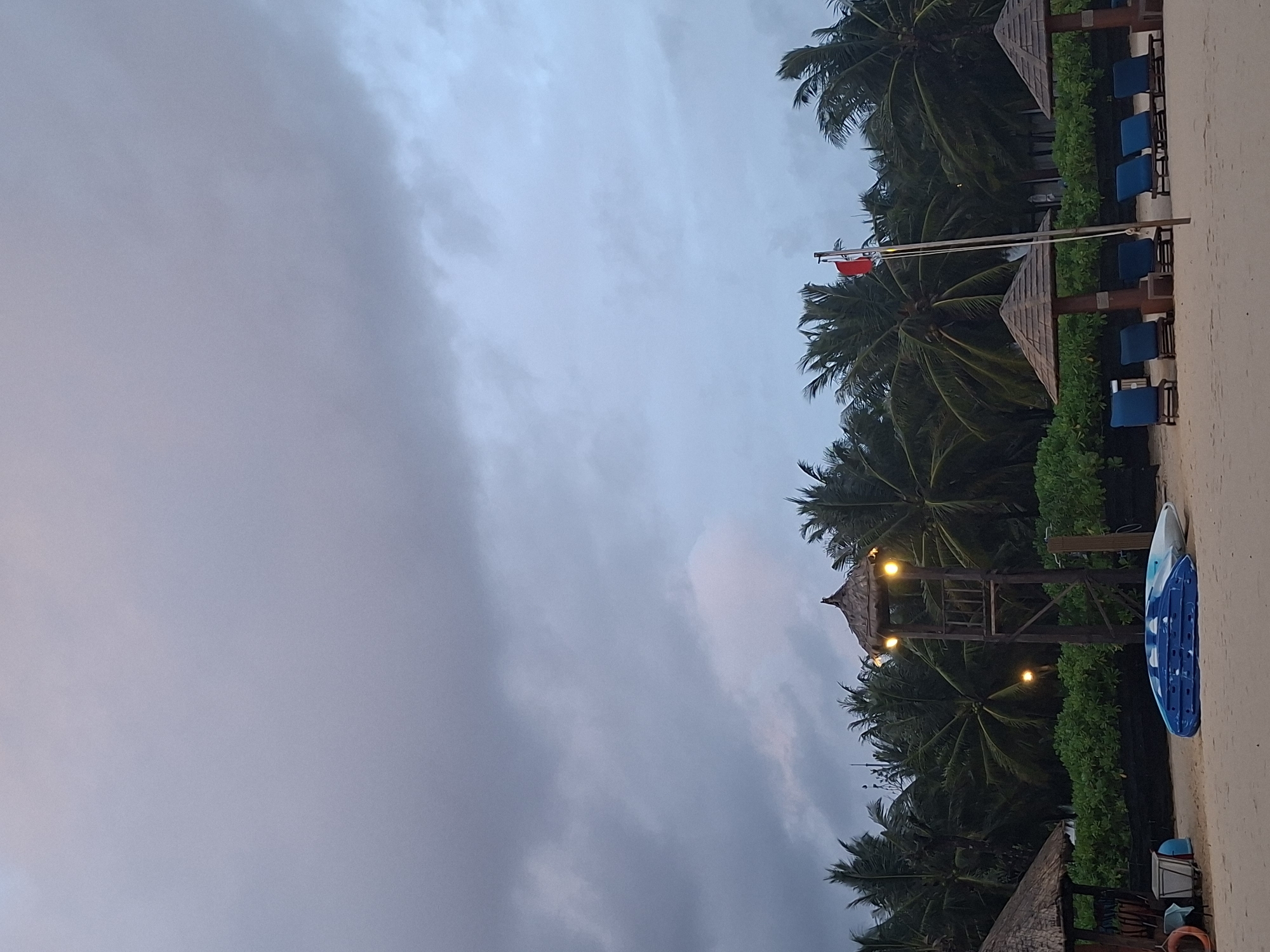 The image depicts a beach scene in Phú Quốc, Vietnam, under a cloudy sky. The foreground is a sandy beach with several lounge chairs and umbrellas, a lifeguard tower, and two kayaks. The background features a dense line of palm trees. A red flag flies from a flagpole near the center of the image. The lighting suggests it is either early morning or late evening, with the sky cast in muted shades of gray and blue. No people are visible in the frame.