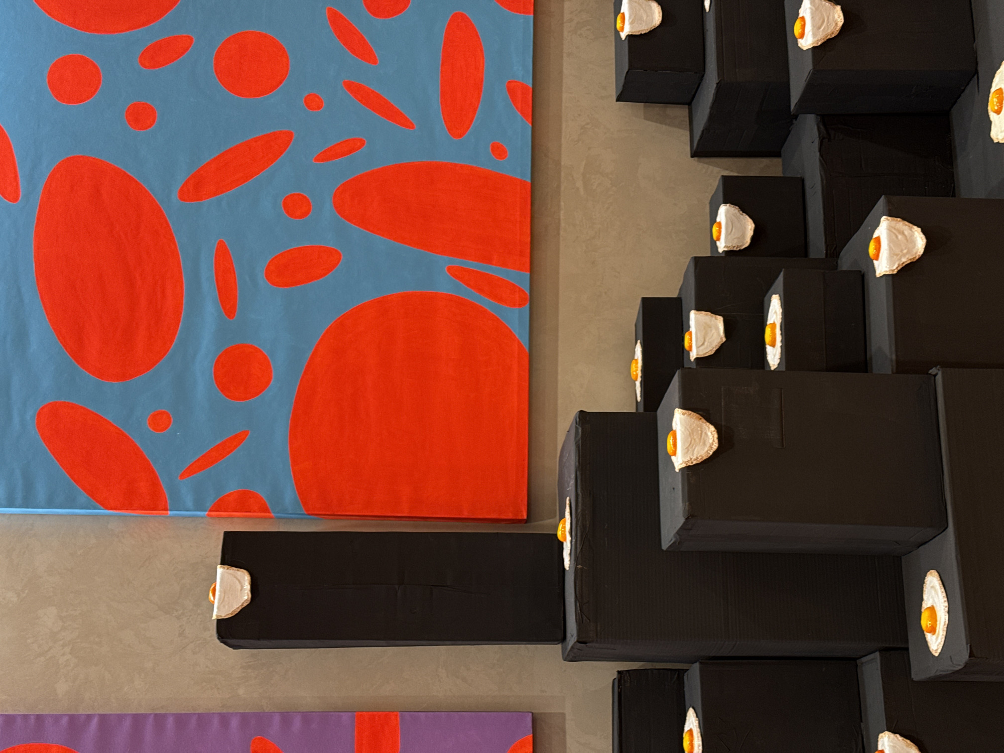 The image captures a vibrant and contrasting art installation, likely within a gallery or exhibition space in Milan, Italy.
On the left, a large canvas displays an abstract painting characterized by numerous red, organic shapes—including circles and ovals of varying sizes—set against a light blue background. The shapes are irregularly scattered, creating a dynamic and fluid pattern that extends beyond the visible edges of the canvas.
To the right, a three-dimensional wall sculpture or installation is prominently featured. It is composed of multiple black, rectangular blocks that are stacked and arranged in a seemingly random yet structured manner, protruding from a neutral, light brownish-grey wall. A distinctive and repetitive element of this installation is the small, realistic-looking fried egg sculptures attached to the faces of many of these black blocks. Each egg exhibits a detailed white and a bright orange yolk, providing a surprising, everyday motif against the stark, geometric backdrop.
A portion of another artwork is visible at the very bottom of the frame, revealing a purple canvas with similar red, organic shapes, suggesting a cohesive artistic theme or series. The overall impression is one of contemporary art that juxtaposes bold colors, abstract patterns, and unexpected sculptural elements. No people are present, and no visible text can be discerned in the image.