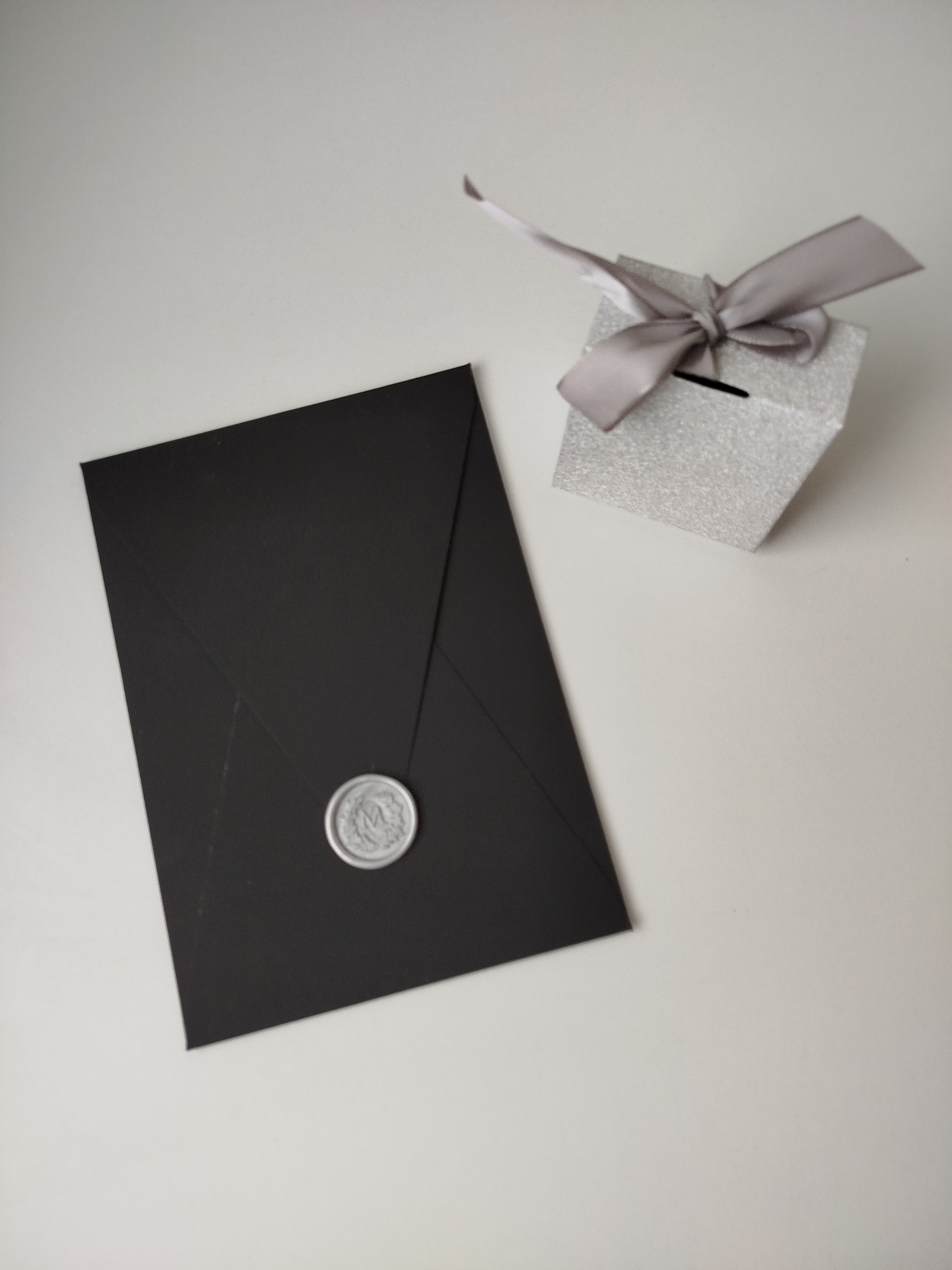A silver glittery gift box with a grey ribbon and bow sits to the right of a black envelope sealed with a silver wax seal. The items are on a white surface. The setting is not identifiable as Varna, Bulgaria.