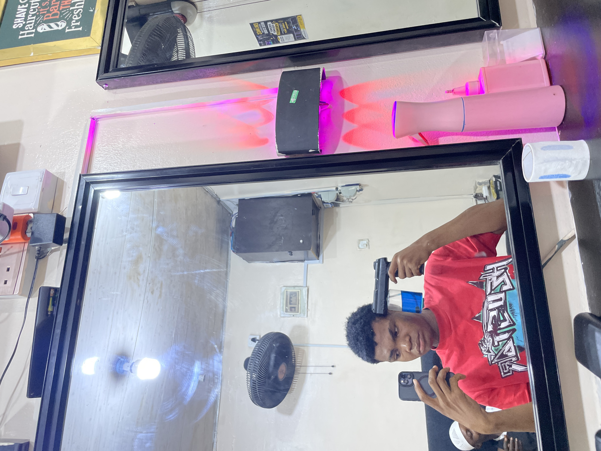 A mirror reflects a young Black man in a red T-shirt holding a black handgun to his head while simultaneously holding a smartphone to take a picture or video. The mirror is mounted on a wall in what appears to be a barber shop in Ado, Nigeria. Reflections in the mirror show additional items such as an air conditioner, an electric fan, and a white baseball cap. Above the mirror are barber shop supplies and another mirror with a sign.
