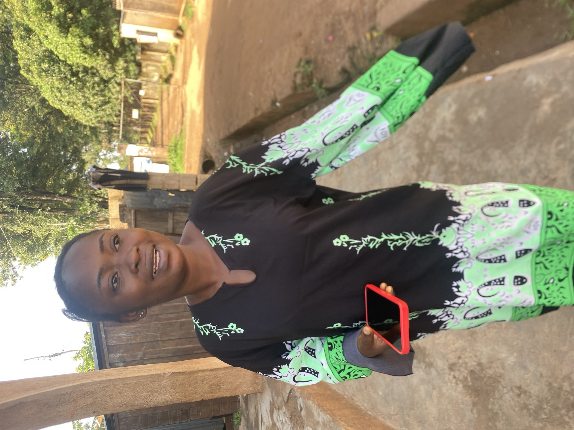 In Bukuru, Nigeria, a young Black woman stands on a paved path outdoors. She is wearing a long-sleeved black dress with a green and white pattern along the sleeves and hem. Her right hand holds a red-cased smartphone. Behind her, a dirt road stretches into the distance, bordered by a fence and trees. The scene appears to be a residential or community area.