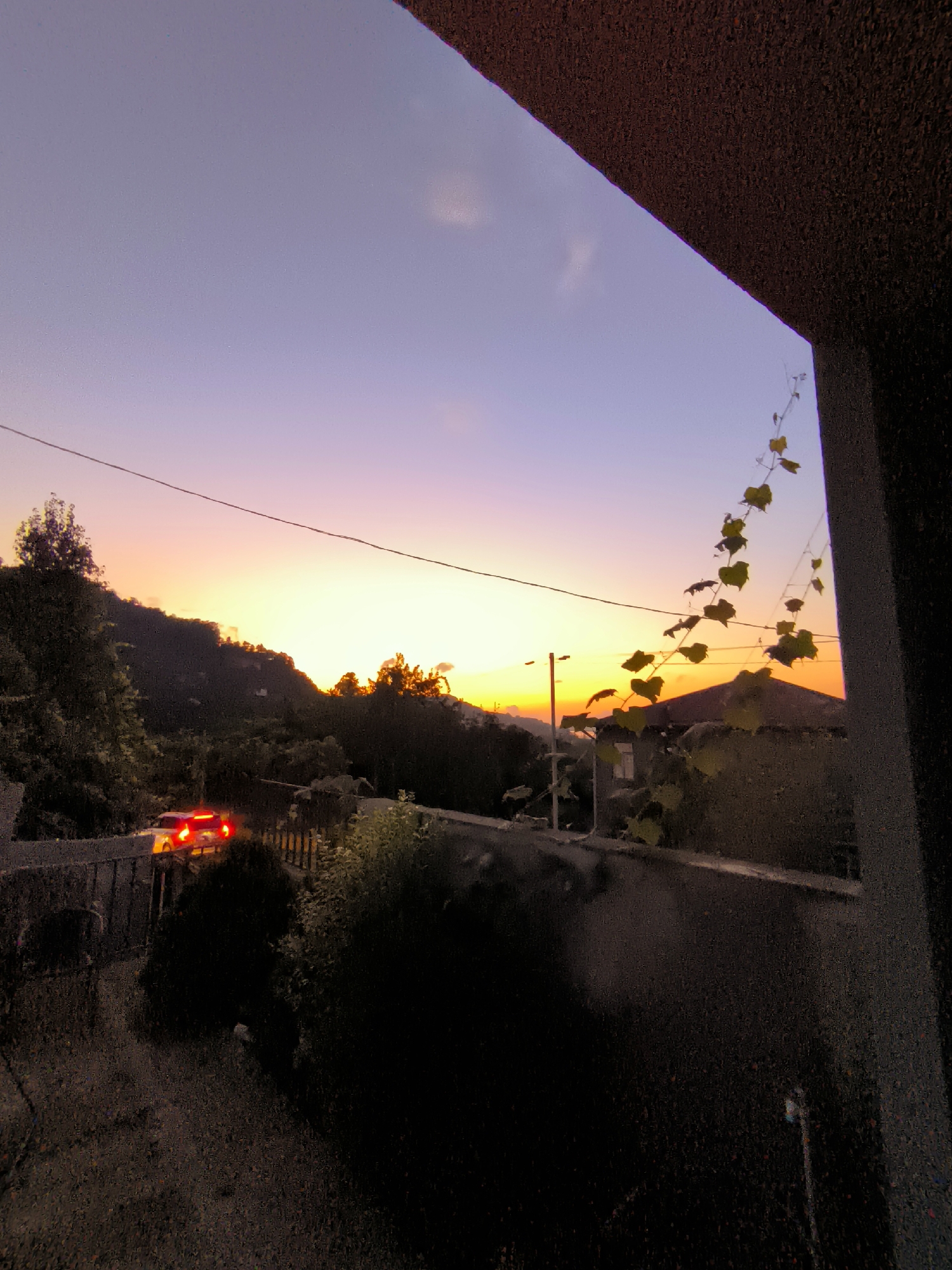 The image captures a sunset scene in an unidentified location. The sky displays a gradient of colors, transitioning from a light purple at the top to a bright orange near the horizon. Silhouetted against the sky are rolling hills covered in dense foliage. A single power line stretches horizontally across the frame, intersecting the sky. In the foreground, on the left, a car with its brake lights illuminated is visible on a road that curves around a hillside. The car appears to be stopped or moving slowly. To the right, a portion of a building's roof is visible, partially obscured by a vine with yellowing leaves that dangles into the frame. The overall impression is one of a serene, natural landscape at dusk, with a touch of human presence indicated by the car and building.