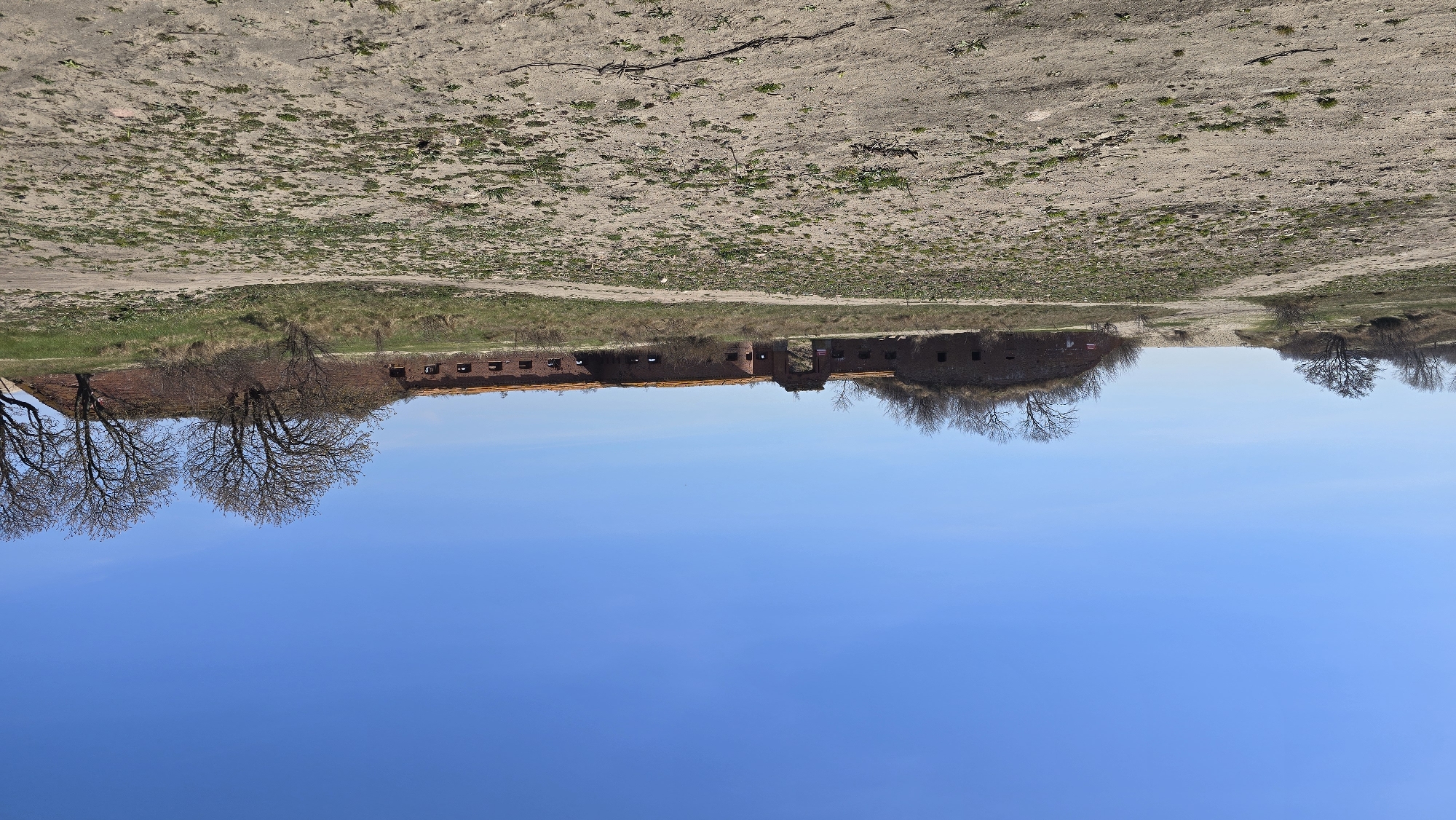 The image displays an upside-down view of a landscape featuring a fort under a clear blue sky. The fort appears to be made of red brick and has a fortified structure with arched windows and doorways. The fort is surrounded by vegetation, including grass and bare trees. The landscape around the fort appears arid, with a mix of bare ground and sparse vegetation. This is the Starotorpednyy Fortress, located in Baltiysk, Russia.