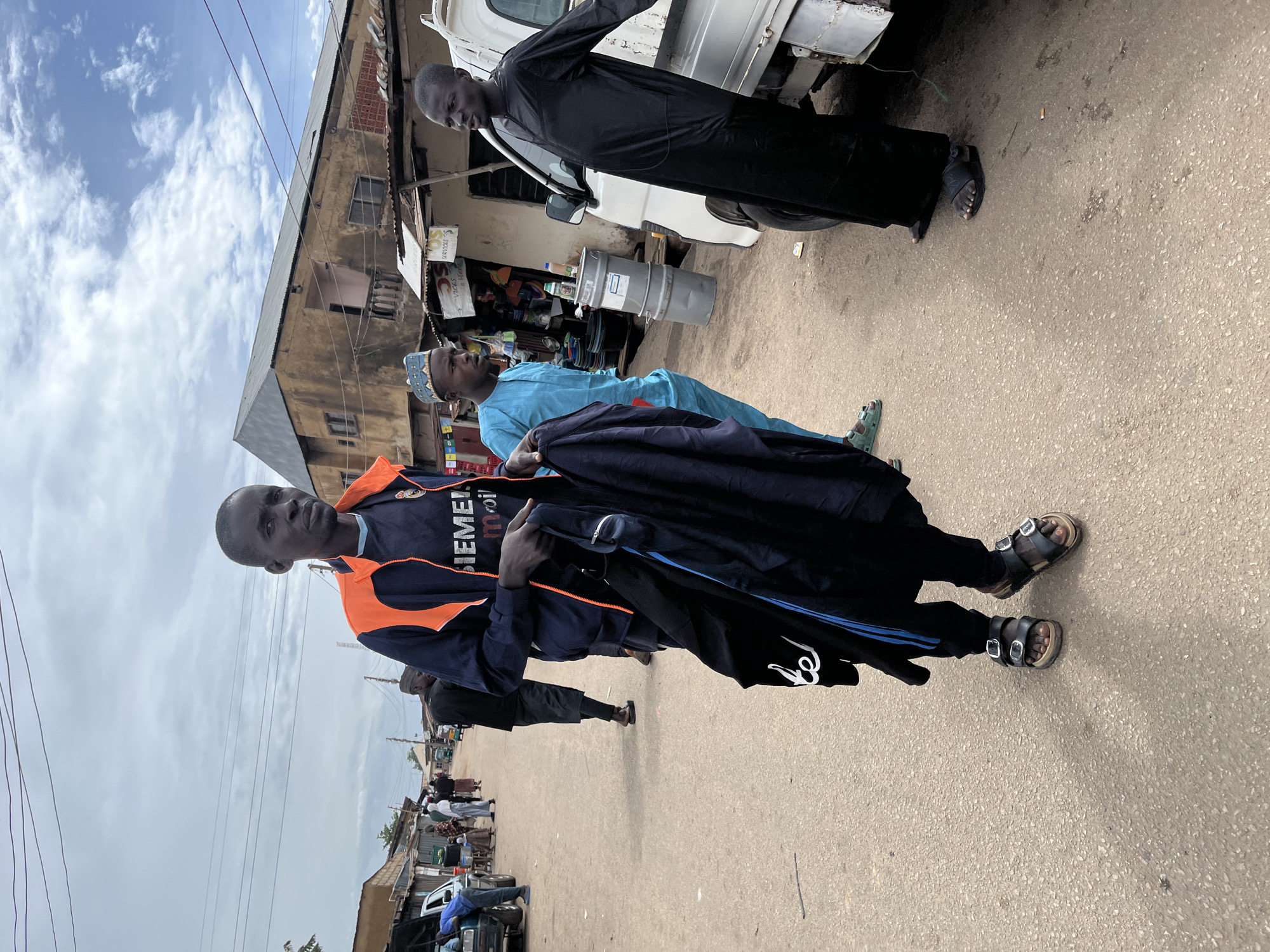 This outdoor scene in Gura Top, Nigeria, depicts several men on a street under an overcast sky. In the foreground, a man stands facing left, wearing a navy blue and orange jacket with "SIEMENS" and "mobil" written on it, dark pants, and black sandals. He holds a dark jacket and what appears to be a pair of dark track pants. His expression is neutral as he looks towards the camera.
Behind him and slightly to the right, another man, dressed in a light blue traditional tunic and matching cap, also holds dark clothing. He looks towards the right side of the frame. Further back, near a white pickup truck, a third man in a black traditional robe stands, looking downwards.
The setting is a street with worn asphalt and scattered debris, including a cigarette butt near the truck. Along the street, single and two-story buildings, appearing somewhat dilapidated, house various small shops or market stalls. One sign in the background clearly shows the word "VICKS." Overhead, utility lines crisscross against the cloudy sky. In the distance, more people and parked vehicles are visible, suggesting a bustling, everyday street environment. The time of day is daylight, indicated by the natural illumination, though the sky is largely cloudy. The overall mood is observational, capturing a moment of daily life.