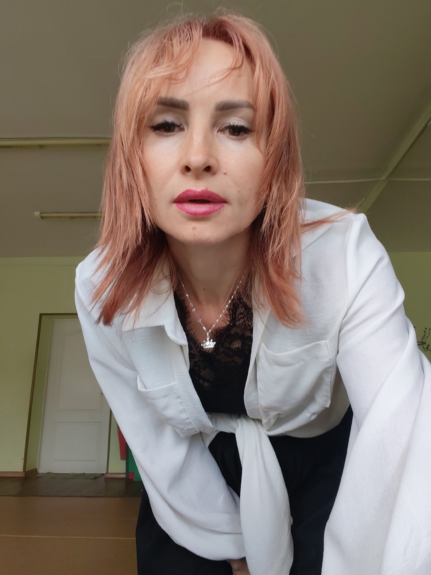 A woman with pink-tinted hair is pictured from the chest up. She is wearing a white, unbuttoned shirt over a black lace top and a silver necklace with a flower pendant. Her makeup includes dark eyeliner and bright pink lipstick. The background shows a cream-colored ceiling with fluorescent lights and a greenish-yellow wall with a white door. The flooring appears to be brown. The overall environment suggests an indoor location, possibly a studio or office space, located in the city of Chaikovsky, Russia.