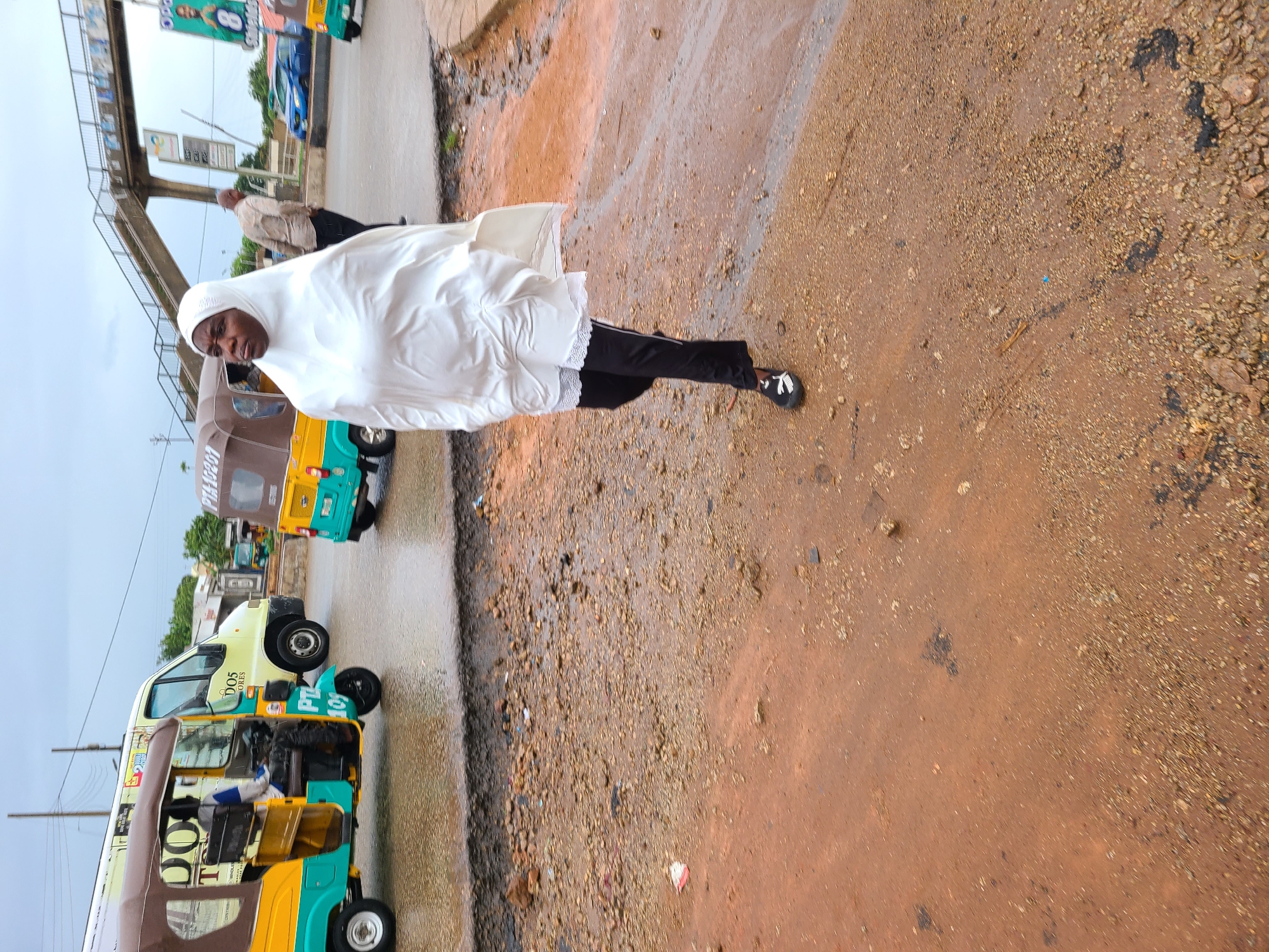 A woman wearing a white hijab and a white abaya with black leggings and black and white sneakers walks on a muddy, unpaved road in Gura Top, Nigeria. She is walking towards the right side of the frame, looking slightly to her left. Her abaya is billowing in the wind. In the background, there are several auto-rickshaws, a white van, and a pedestrian overpass. The road appears to be wet and muddy, indicating recent rainfall. The sky is overcast.