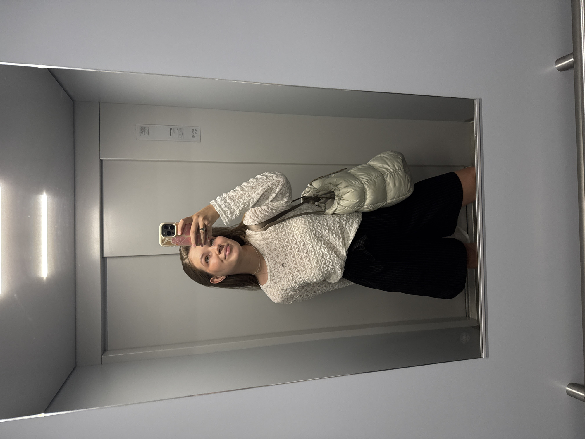 A young woman with light brown hair takes a mirror selfie in an elevator. She is wearing a white, textured long-sleeved shirt and black shorts. A light-colored puffer bag hangs from her right shoulder. The elevator has grey walls and automatic doors. The overall lighting is bright and even, likely from the elevator's ceiling light. The scene is clean and utilitarian. The location is identified as an elevator in Vozdovac Urban Municipality, Serbia.