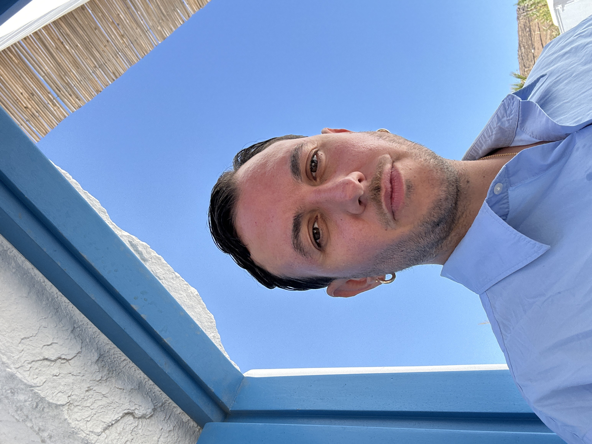 In Ano Mera, Greece, a man with dark hair and a light stubble is pictured in a close-up, self-taken photograph. He is wearing a light blue button-down shirt and a gold chain. He has a gold hoop earring in his left ear. The man is framed by architectural elements: on the left, a textured white surface meets a vibrant blue beam, and on the right, a thatched roof structure is visible against a clear blue sky. The lighting suggests it is daytime, and the overall scene evokes a relaxed, possibly vacation atmosphere.