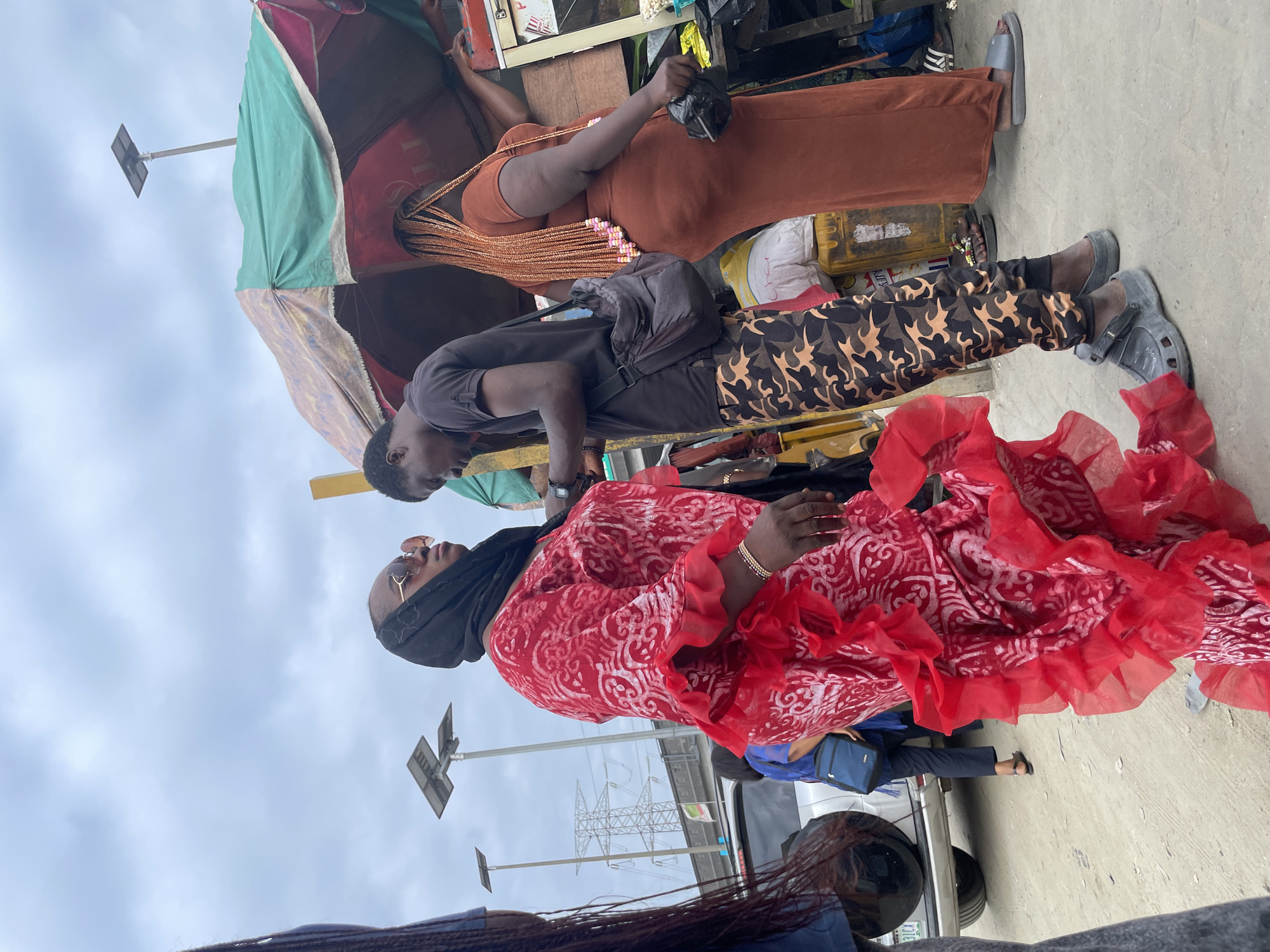 In Lekki, Nigeria, on an overcast day, a group of people are gathered near market stalls. A woman in a vibrant red dress with ruffled sleeves and a black headscarf is in the foreground, facing left and looking away. She wears sunglasses and a bracelet. Next to her, a man in a black t-shirt and camouflage pants stands and looks down at his phone. To the right, another woman in a long, rust-colored dress with braided blonde hair decorated with pink and white beads is leaning on a market stall. The stall is covered by a multi-colored awning. In the background, a white SUV and a power pylon are visible. The ground is unpaved, and the overall atmosphere suggests a busy outdoor market.