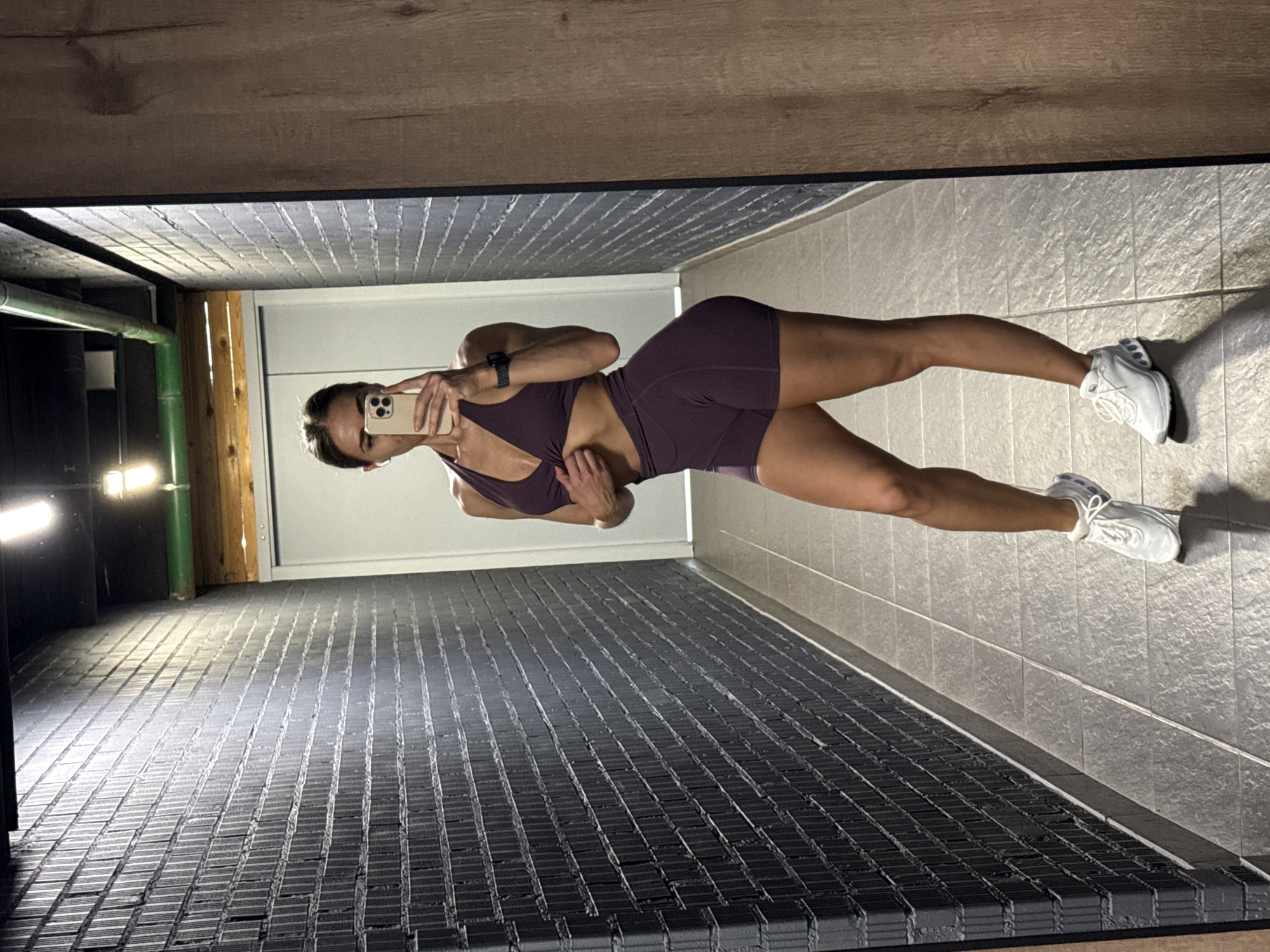 The media file is a vertically oriented, full-body mirror selfie taken by a woman in athletic wear. She is wearing a dark purple sports bra and matching shorts. She is holding a smartphone in her right hand, angled to capture the reflection. Her left hand is resting on her hip. She has on white athletic shoes.
The setting appears to be an indoor space, possibly a gym or a locker room, given the flooring and the presence of what looks like ventilation pipes and wooden beams. The floor is covered with textured, dark grey tiles that appear to be industrial or gym-style mats. The walls are tiled with lighter grey square tiles. The lighting suggests it might be indoors, possibly with artificial lights, as evidenced by the distinct shadows and the bright illumination in the background. The overall tone is neutral and functional. There is no visible text in the image. The woman's posture and attire suggest she is engaged in fitness activities or has just completed a workout.