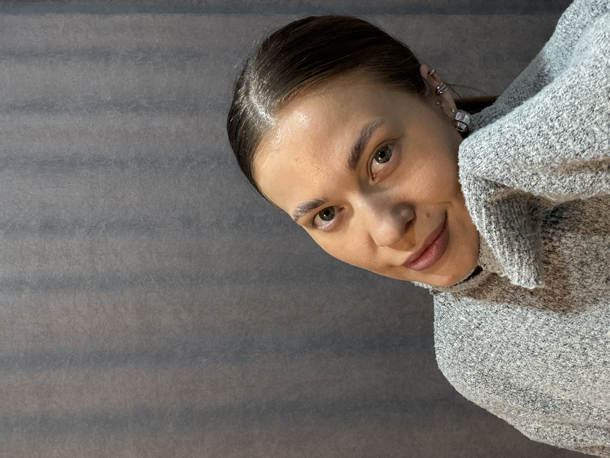 An overhead, close-up portrait captures a young woman looking directly into the camera with a gentle, engaging expression. Her dark hair is neatly pulled back, revealing her forehead and ears. She has green-brown eyes and a soft smile on her lips, enhanced by subtle, natural-looking makeup. On her visible left ear, she wears several silver-toned earrings or ear cuffs, including a prominent piece on the lobe and multiple smaller hoops higher up. She is dressed in a light grey, textured, possibly knitted sweater with a high neckline, which partially covers her chin.
The image is taken from a low angle, looking up at the subject, suggesting a selfie or a portrait from a unique perspective. The background consists of a uniform grey surface with faint horizontal darker bands, indicative of an indoor setting, possibly a wall or ceiling. There are no explicit cues for the time of day or weather conditions, and no visible text. The context for this image is Chernushka, Russia.