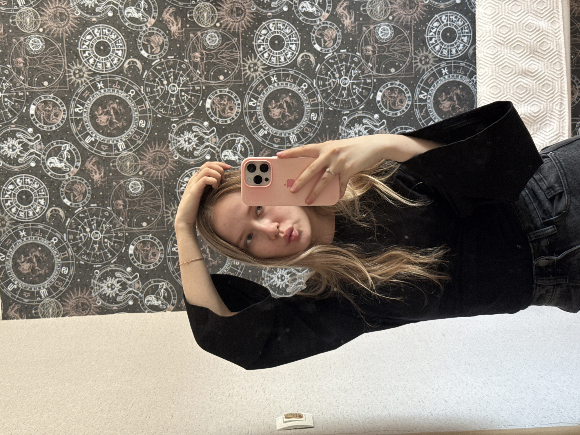 A person with long blonde hair is taking a selfie in a mirror. The person is wearing a black long-sleeved shirt and black jeans. They are holding a pink iPhone in their right hand. The background consists of patterned wallpaper featuring astrological symbols and a white textured surface. The photo was not taken in Zhigalki, Russia.