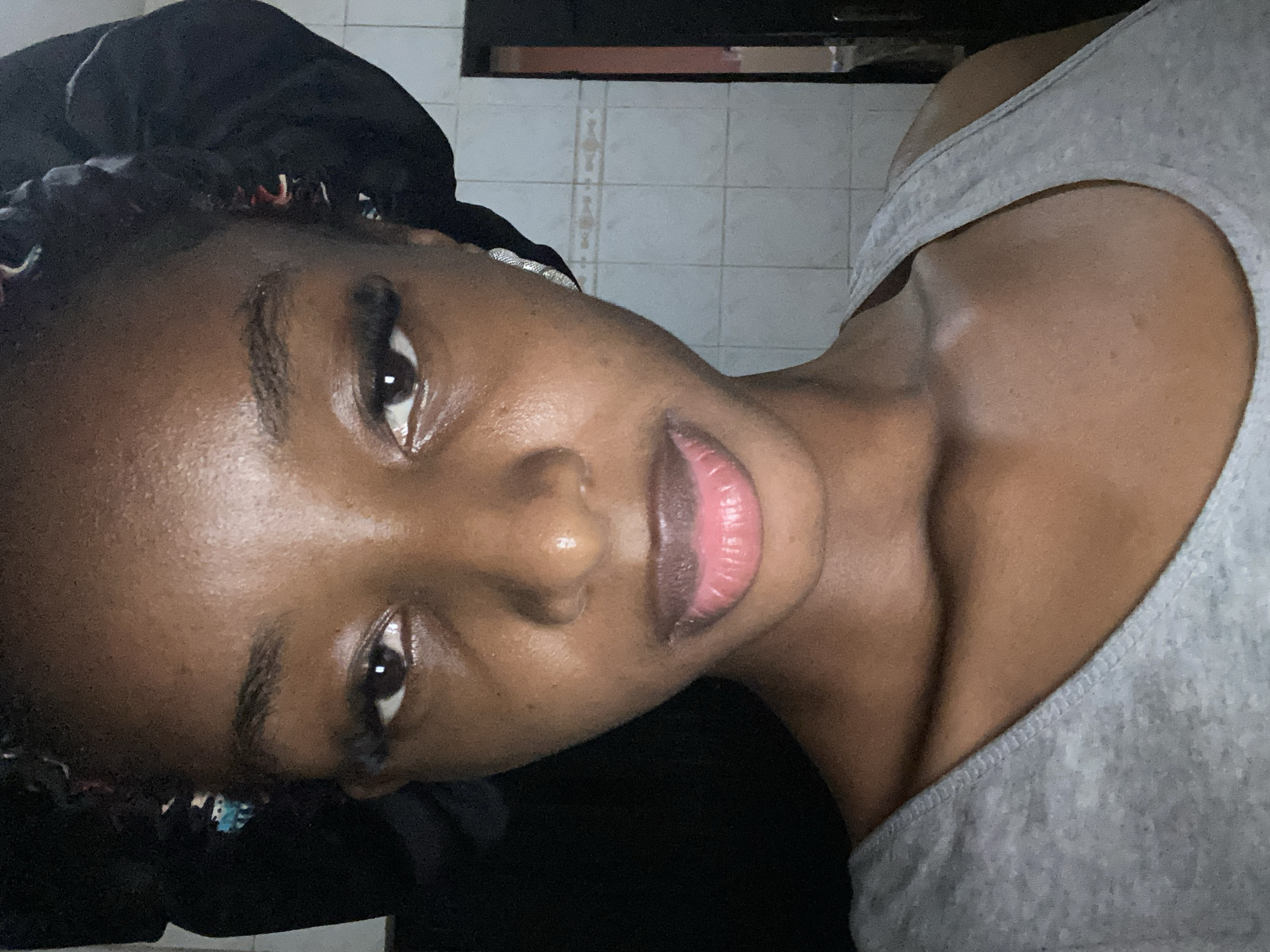 This is a close-up, portrait-style image of a young Black woman. She is wearing a light grey tank top and a black satin bonnet. The image is oriented sideways. Her face is turned towards the camera, with her eyes looking slightly to the side. Her lips are slightly parted, revealing a hint of pink. The lighting in the image is soft, and her skin appears to have a dewy sheen. The background is out of focus, but appears to be a tiled wall, suggesting an indoor setting. There is no visible text in the image. The image conveys a sense of intimacy and self-reflection. The subject's relaxed posture and soft gaze suggest a moment of calm or personal time. The overall mood is subdued and natural. The time of day is not explicitly indicated, but the lighting might suggest indoor lighting. The location context of Jos, Nigeria, does not provide specific visual cues within the image itself.