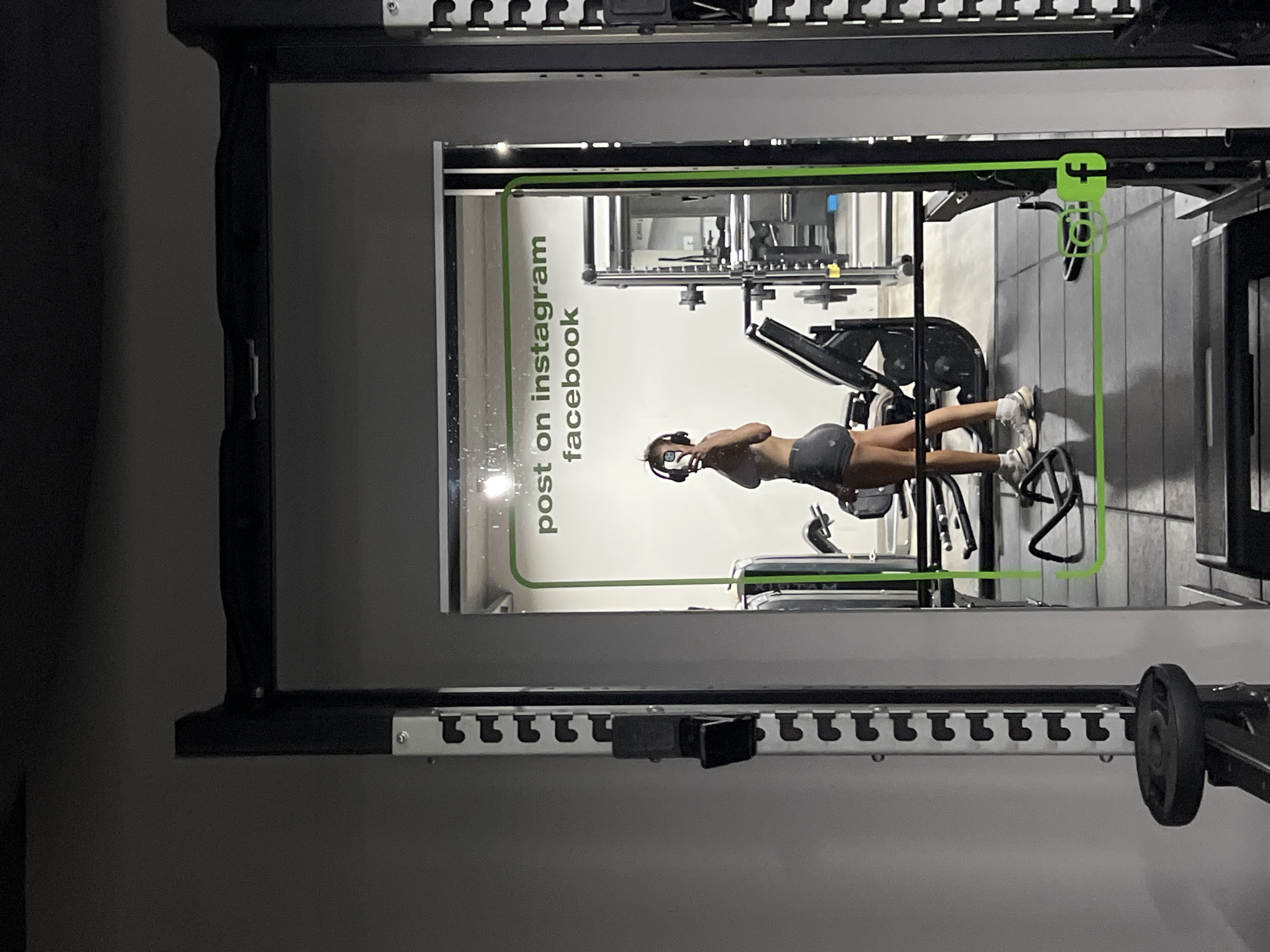 A young woman is taking a mirror selfie in a gym. She is wearing headphones and workout attire, consisting of a sports bra and shorts. The gym equipment, including weight racks and machines, is visible in the background. The mirror has a green border with text that reads "post on instagram facebook" and a Facebook logo. The woman's reflection is the primary subject. The scene is indoors, in a modern gym environment. The location of Mühldorf a. Inn, Germany is not identifiable from the image.