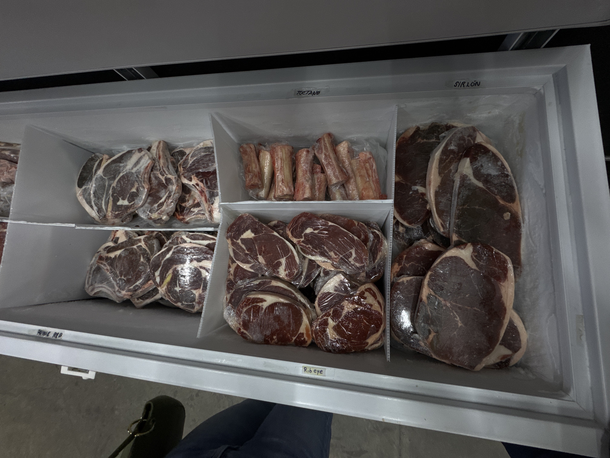 A top-down shot shows a freezer filled with various cuts of meat, likely within a butcher shop or grocery store in Puebla, Mexico. The freezer is divided into sections labeled "Prime Rib," "Tuetano" (bone marrow), "Rib eye," and "Sirloin." Each section contains packages of the corresponding cut of meat, individually wrapped in clear plastic. A person wearing blue jeans is partially visible at the bottom of the frame, along with a green bag. The freezer is gray or white in color.