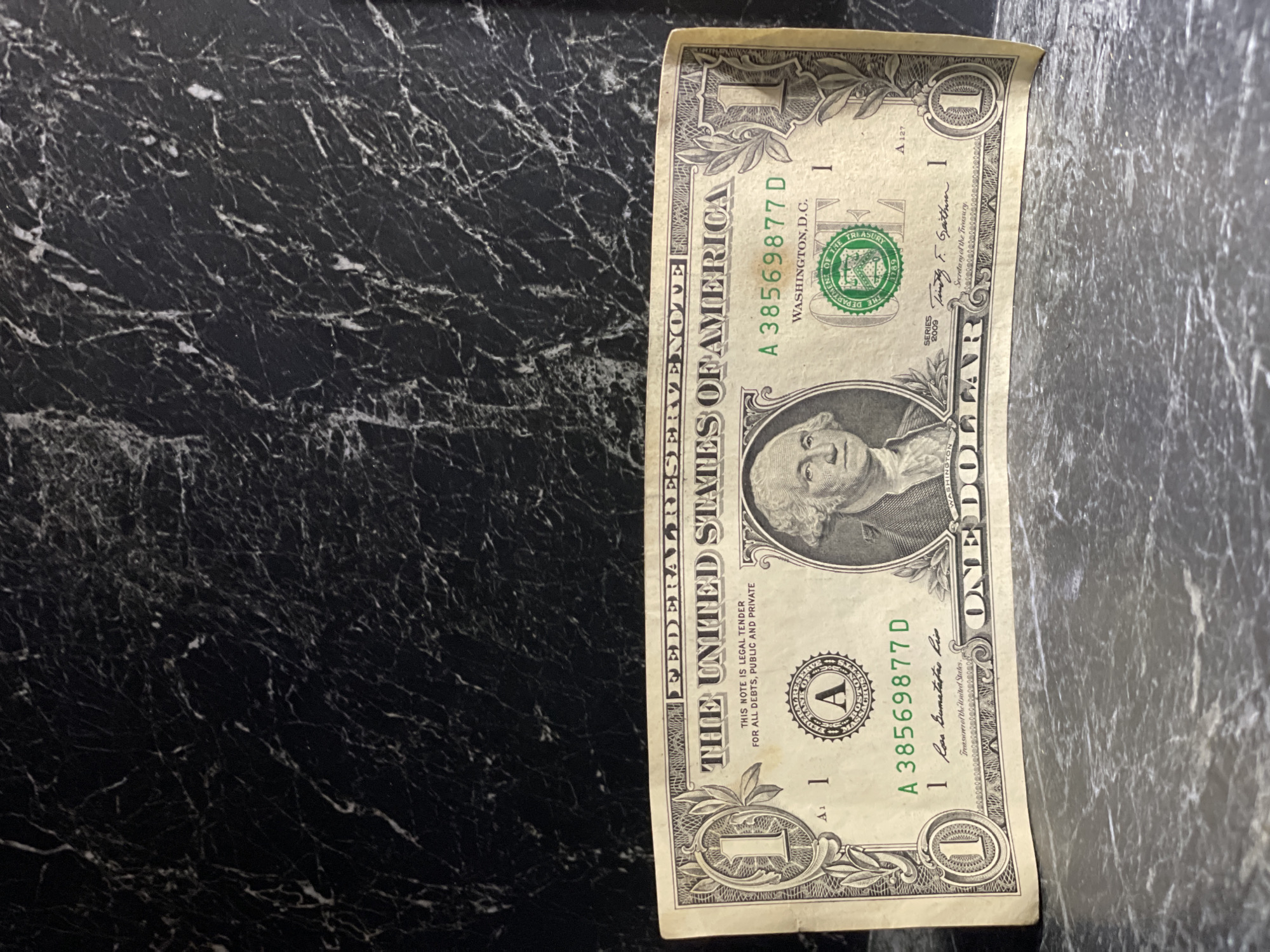 An overhead shot, rotated 90 degrees clockwise, captures a single US one-dollar bill resting on a dark countertop with distinctive white marble-like veining. The bill appears slightly worn and creased, lying vertically on the right side of the frame. The background surface occupies the majority of the image, showcasing its intricate, chaotic white patterns against the deep black.
The dollar bill prominently displays the portrait of George Washington. Visible text includes "THE UNITED STATES OF AMERICA," "FEDERAL RESERVE NOTE," and "ONE DOLLAR." The serial number "A 38569877 D" is clearly seen twice, along with "WASHINGTON, D.C." and "SERIES 2009." The phrases "THIS NOTE IS LEGAL TENDER FOR ALL DEBTS, PUBLIC AND PRIVATE" and "Secretary of the Treasury" (with the signature of Timothy F. Geithner) and "Treasurer of the United States" (with the signature of Rosa Gumataotao Rios) are also legible. A green seal of the Department of the Treasury (1789) and a black Federal Reserve Bank Seal with the letter "A" (indicating the Federal Reserve Bank of Boston) are present. The number "1" representing the denomination is visible in multiple locations on the bill. The image suggests this US currency is being observed in Kazan, Russia.