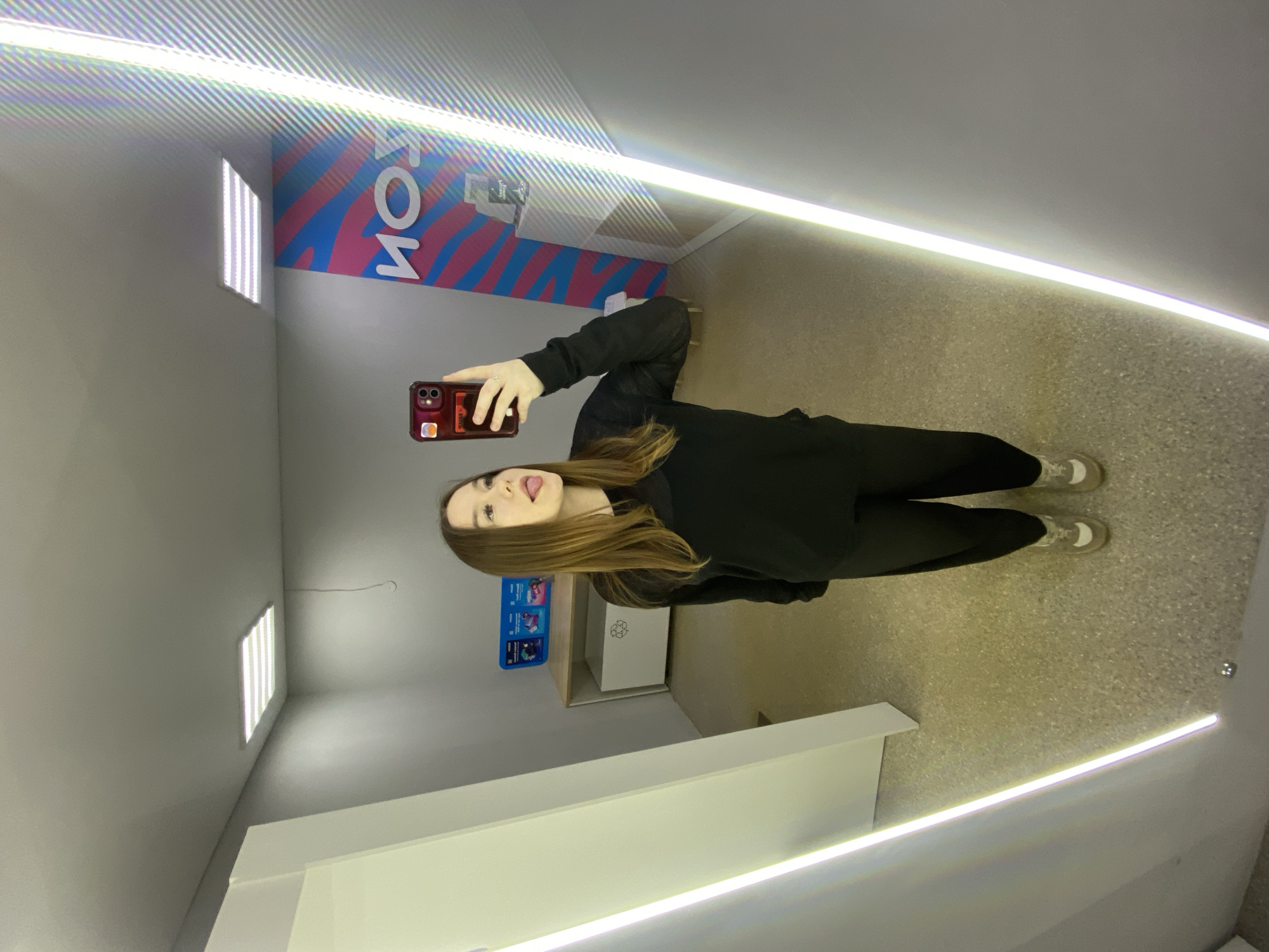 This media file captures a young woman taking a mirror selfie in a modern, brightly lit indoor space, likely located in Chaikovsky, Russia. The image is oriented vertically, with the woman appearing on her side, though she is standing upright in the actual scene.
The central subject is a young woman with long, straight brown hair, dressed casually in a black hoodie and black pants, paired with white sneakers. She is captured in a playful moment, sticking out her tongue while looking directly into the smartphone she holds up to take the photo. Her phone is encased in a red cover adorned with a few stickers.
The setting is characterized by its contemporary design and abundant artificial lighting. Bright, linear LED lights illuminate the space, running along the edges of the walls and ceiling, creating striking reflections on the various surfaces. The walls are predominantly light grey, and the floor is a light-colored, speckled concrete or similar industrial material. In the background, a wall features a vibrant graphic design with blue, pink, and white patterns. Reflected on this wall is the text "ТОЙ", which, when viewed in its correct orientation, suggests it might be part of a larger word such as "АКРОН" (ACRON). Below this graphic, there's a low, light-colored counter or shelf, and a white recycling bin marked with the universal recycling symbol. Another rectangular light panel is visible on a far wall.
The activity is a self-portrait taken via a mirror, highlighting a casual and lighthearted mood through the woman's playful expression. The scene is brightly lit by artificial sources, making the time of day and external weather irrelevant. The combination of modern lighting, reflective surfaces, and functional elements like the recycling bin suggests a public, commercial, or possibly an exhibition space.