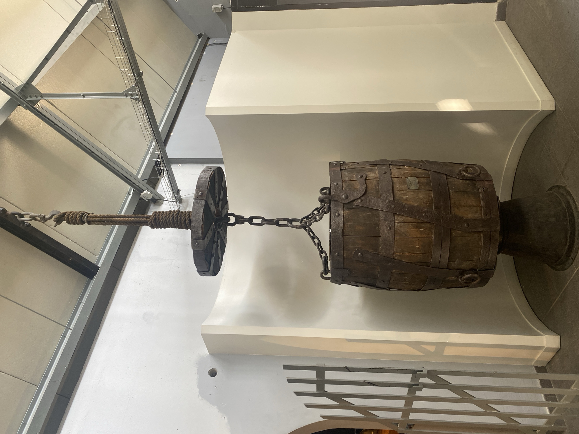 The image shows a medieval torture device on display in what appears to be a museum or historical exhibition. The device consists of a wooden barrel bound with metal straps, suspended by chains from a pulley system. The pulley system includes a wheel and rope, which are attached to a metal frame. The device is mounted on a white pedestal with curved supports. The background features white walls and a ceiling with visible metal supports. The location is suggested to be in La Motte-d'Aveillans, France. There are no people visible in the image. The object's primary function is to serve as a display, likely aiming to educate viewers about historical torture methods.