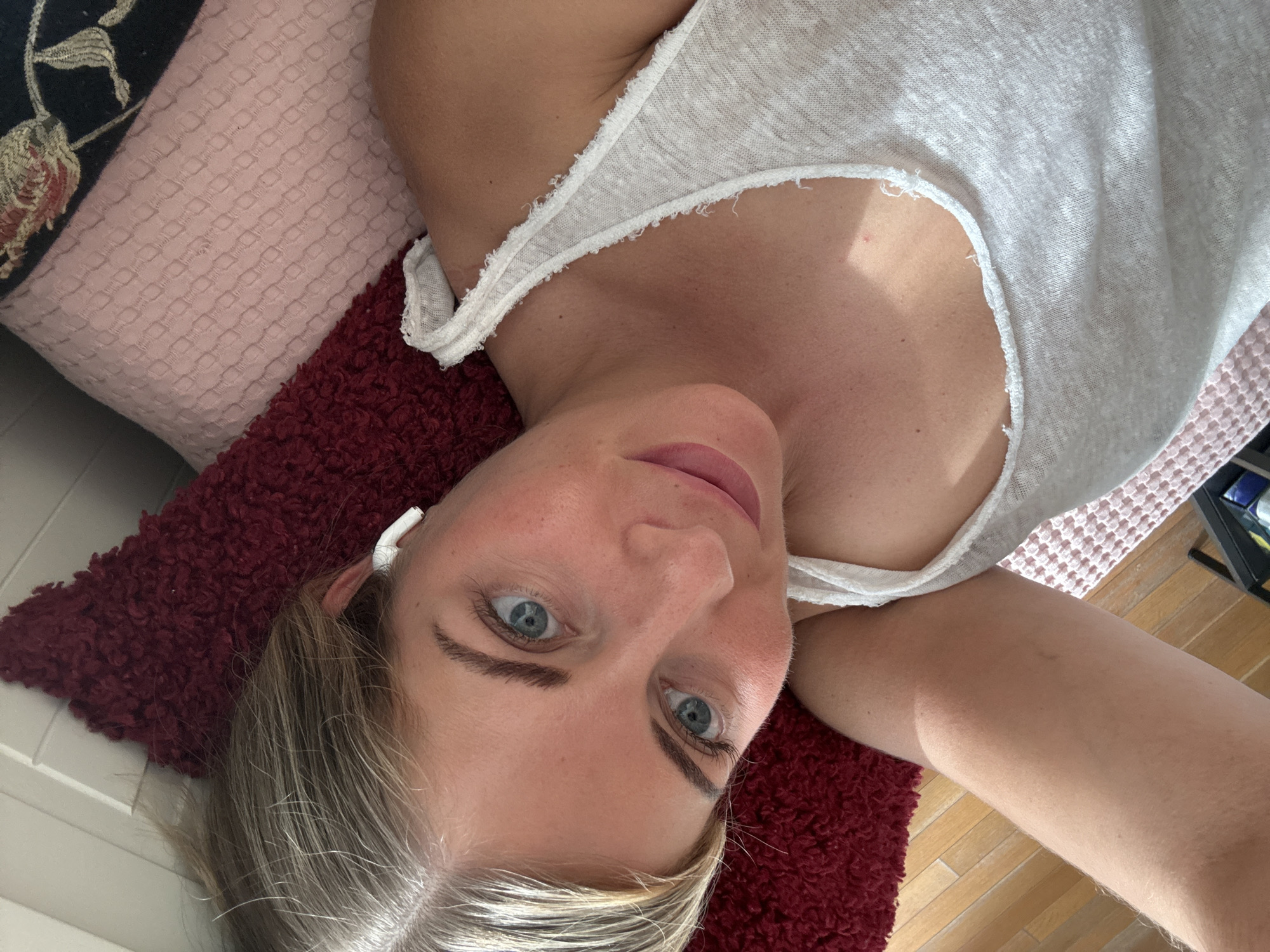 A fair-skinned woman with blonde hair and blue eyes is lying on her back, looking up at the camera. She is wearing a white tank top. Her head rests on a red, textured pillow, which is placed on a bed that is covered in a pink textured sheet. Behind the woman’s head are several decorative pillows, one of which is floral. The image was likely taken indoors. The location in Paris, France is not identifiable from the image.