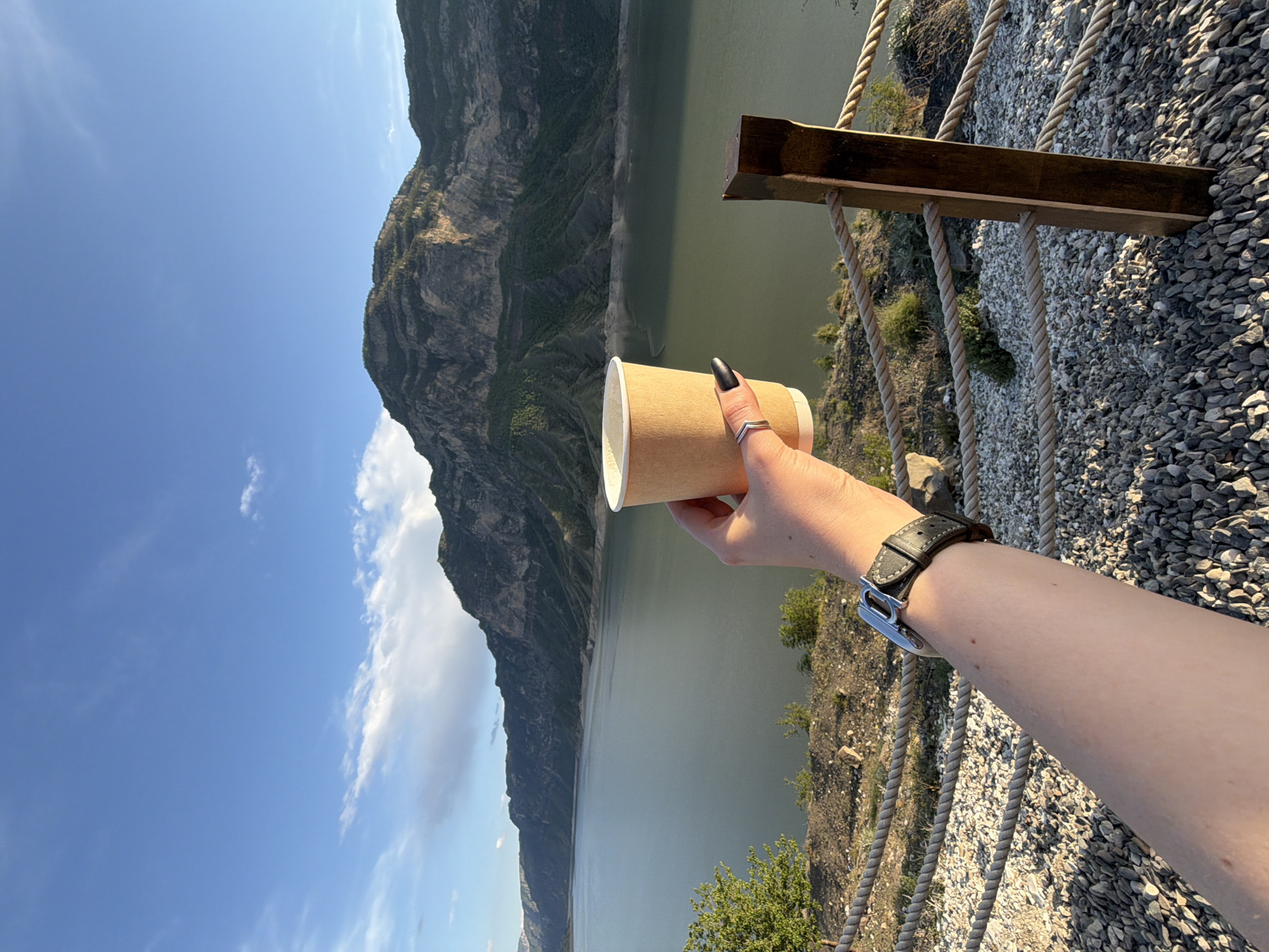 A view of a person's hand holding a coffee cup over a mountain lake. The hand is fair-skinned with black nail polish and a silver ring. A black leather watch is on the wrist. The background features a mountain range, a lake, and a fence. The location is Unknown, Unknown.