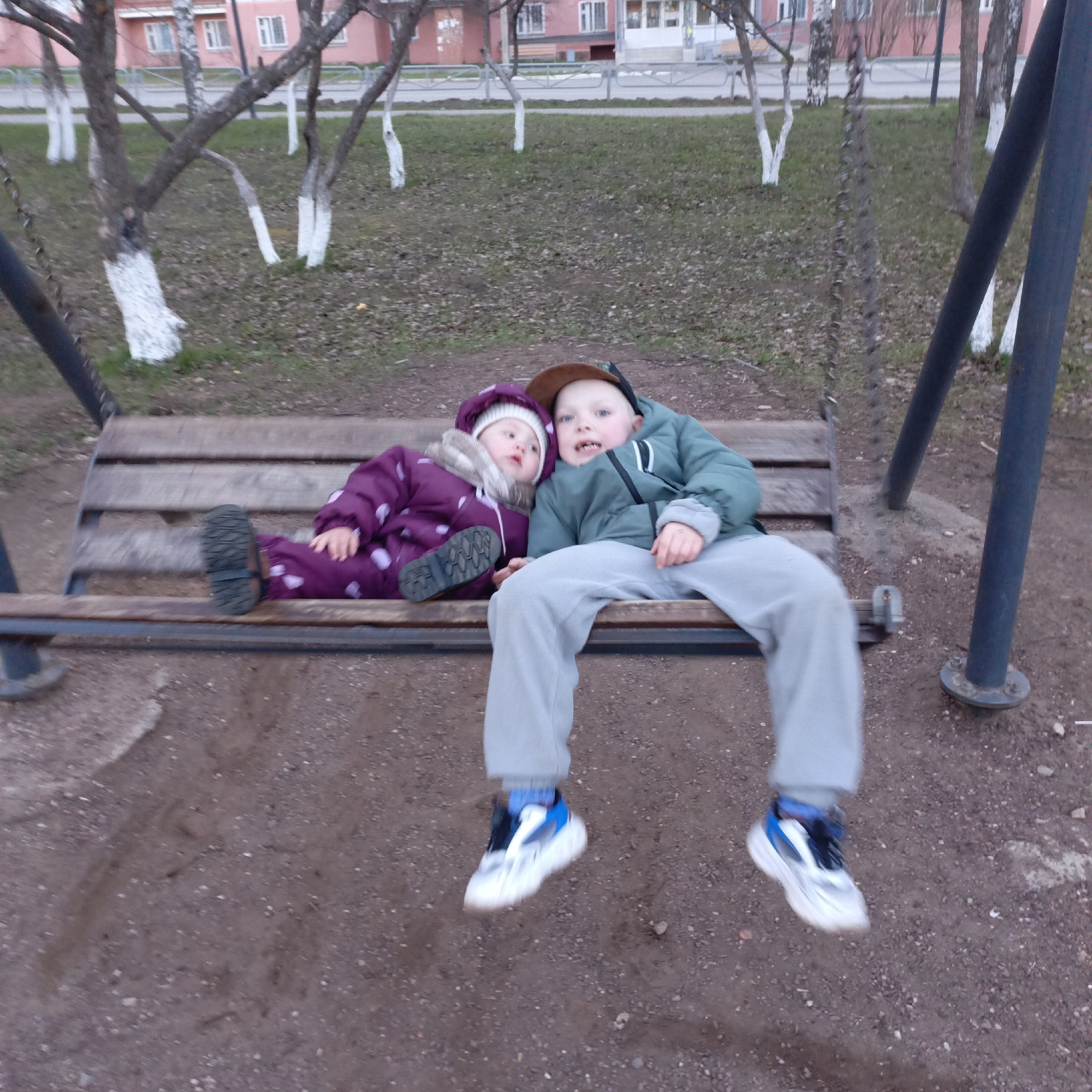 This image captures two young children, a boy and a girl, sitting together on a wooden swing bench in an outdoor park or playground setting. The boy, positioned on the right, wears a green puffer jacket, grey sweatpants, a brown baseball cap, and white, blue, and black sneakers. He looks towards the camera with a slight smile. The girl, on the left, is clad in a warm purple, polka-dotted snowsuit or overall, and a white knitted hat, with black boots visible. She leans her head on the boy's shoulder, appearing relaxed or perhaps sleepy.
The swing itself consists of a slatted wooden bench suspended by dark metal chains from a sturdy metal frame. The ground beneath the swing is composed of dirt and small gravel. In the background, several deciduous trees with distinctive white-painted trunks are scattered across a grassy area mixed with fallen leaves. Behind the trees, residential buildings with light-colored windows and reddish-pink facades are visible, suggesting an urban or suburban environment. A low fence or barrier separates the grassy area from what appears to be a paved path or road.
The overall lighting suggests it is either late afternoon or early evening, or an overcast day, as the light is soft and diffused with no strong shadows. The children's warm attire indicates cool weather. The blur around the boy's feet implies that the swing is in motion, suggesting they are actively swinging or have just finished. There is no visible text in the image. The scene conveys a sense of youthful companionship and calm enjoyment in an outdoor play area.
