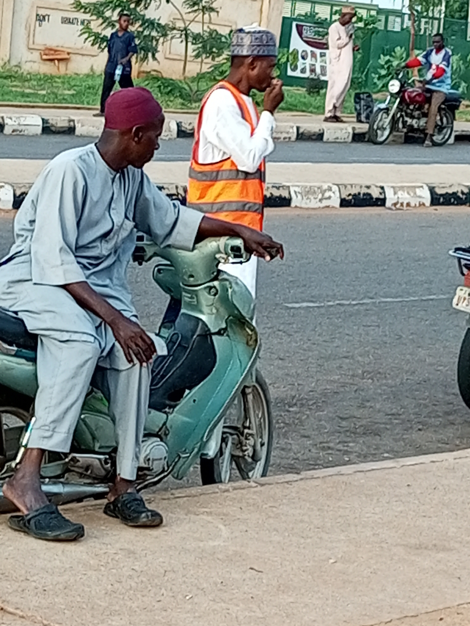 In Gombe, Nigeria, an individual wearing a maroon cap and a light gray traditional outfit is seated on a light green motorcycle. The motorcycle is positioned on the edge of a paved road, near a concrete curb. The person's left foot is resting on the ground with a black sandal, while the right foot is on the footrest of the motorcycle. Another person, wearing an orange reflective vest over a white shirt and a patterned cap, stands behind the motorcycle, facing left and appearing to be eating. In the background, a person in a light-colored robe and cap stands by a green fence, and further in the background, a person in a blue outfit stands near a wall with writing on it. Two motorcycles are visible in the scene, one being ridden by a person wearing a blue and red shirt and brown pants. The environment suggests an urban setting with a road, curb, and a building facade.