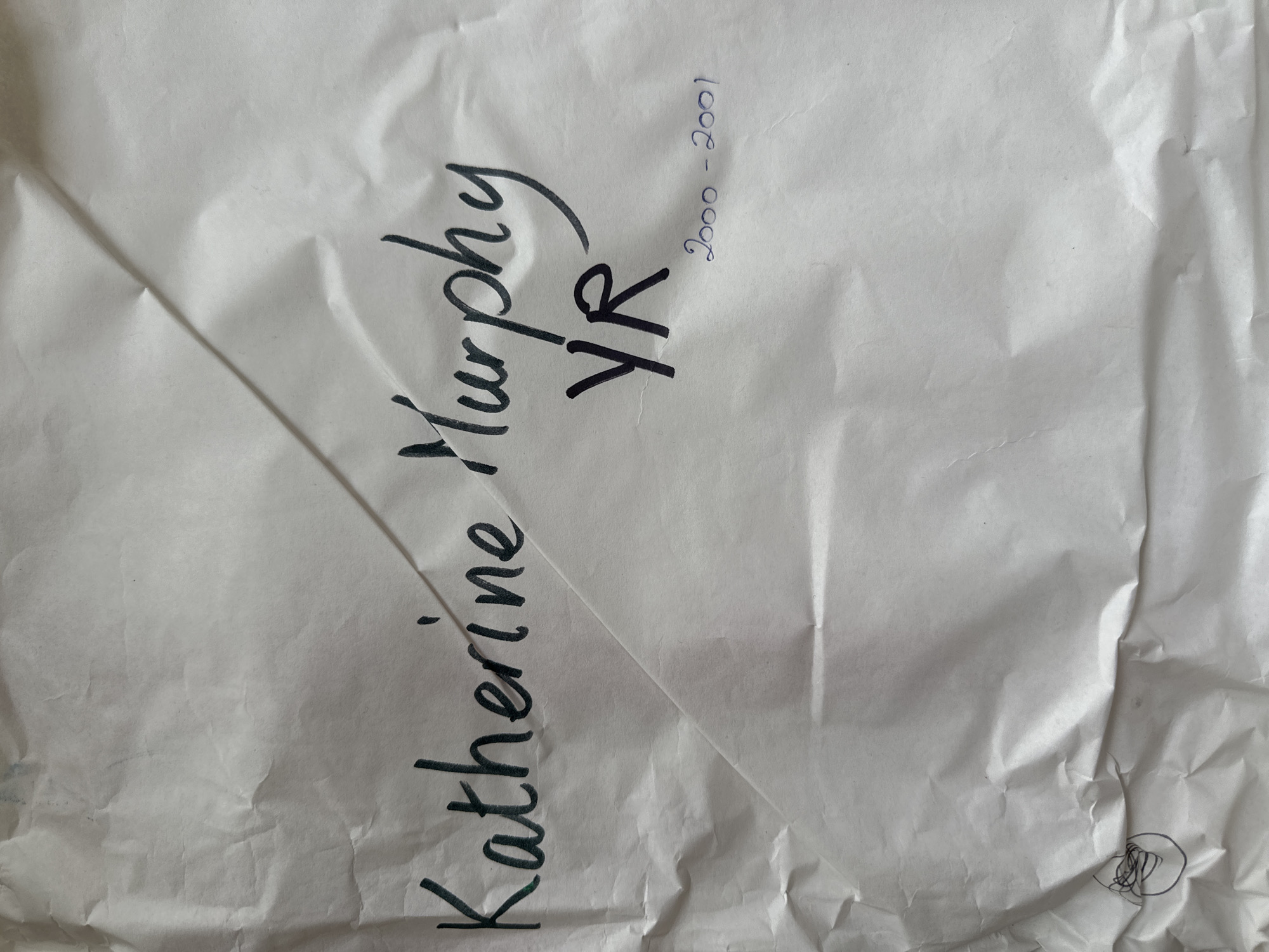 A single sheet of heavily creased, off-white paper is depicted from an overhead perspective. The paper exhibits numerous folds and wrinkles across its entire surface, suggesting prior handling or storage. Prominently centered in the upper-middle section, the name "Katherine Murphy" is handwritten in dark, likely black or dark green, ink with a cursive-like style. Below "Murphy" and slightly to the right, the letters "YR" are written in larger, bolded dark ink. Further below and to the right of "YR," the date range "2000 - 2001" is inscribed in smaller, light blue ink. In the bottom-left corner of the paper, a small, dark, irregular circular scribble is present. No individuals or additional environmental elements are visible. The physical location of this scene in the city of Wealden, United Kingdom, cannot be determined from the visual information presented.