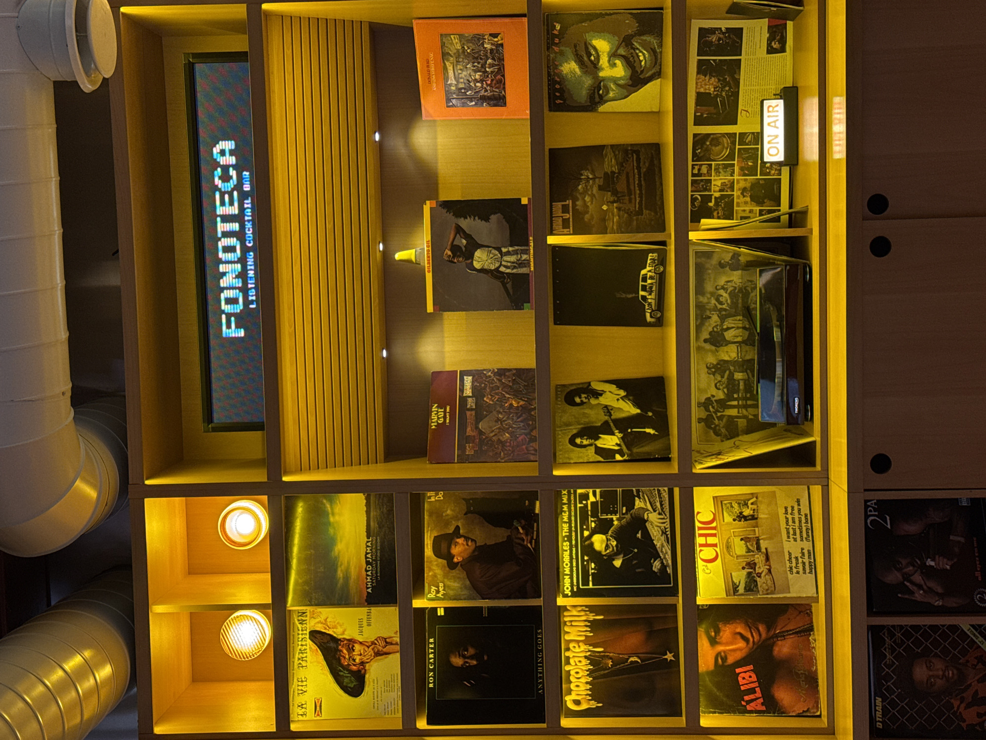 The image shows a wooden shelving unit labeled "FONOTECA LISTENING COCKTAIL BAR" with vinyl records arranged in the cubbies. Visible record sleeves include "LA VIE PARISIENN," "AHMAD JAMAL Saturday Morning," "RON CARTER Anything Goes," "Chocolate Milk," "ALIBI," "GCHIC," "MARVIN GAYE I WANT YOU," "GILBERTO GIL," and "DONALD ISR." An "ON AIR" sign is illuminated on one of the shelves. The location is likely a bar in Yerevan, Armenia. White ductwork is visible on the left side of the frame.