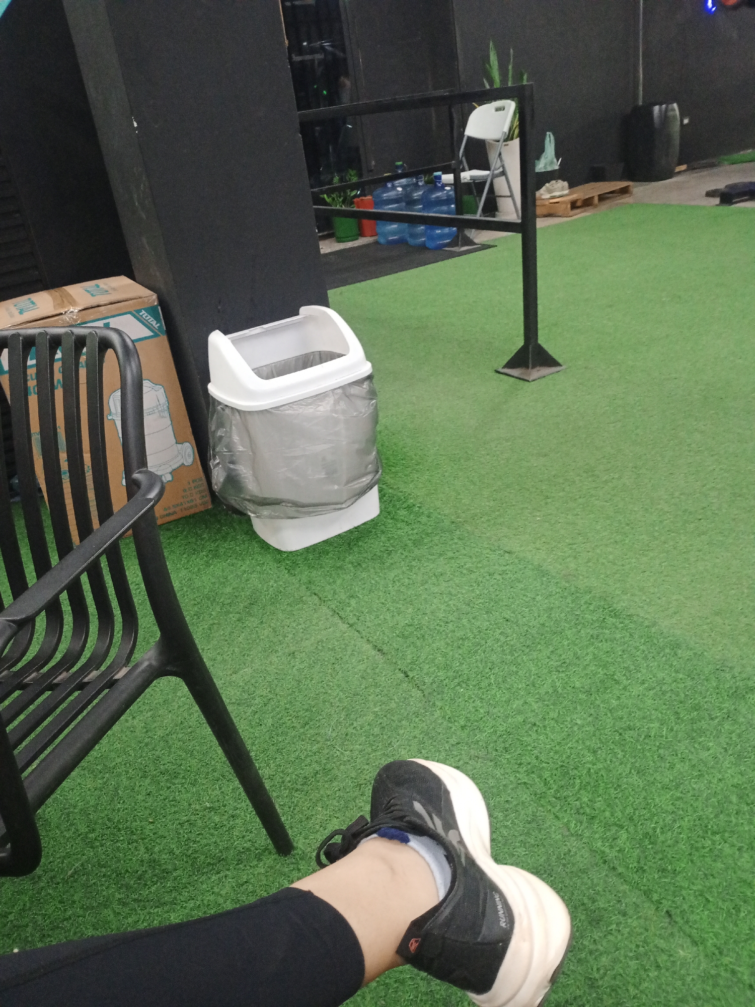 A person is sitting on a black metal chair on green artificial turf. The person is wearing black leggings and a black and white sneaker on one foot. Beside the chair is a white trash can with a gray liner. In the background is a large cardboard box with the word "TOTAL" printed on it. Other gym equipment and a white plastic chair can be seen further back. The photo was taken in a gym or fitness center in Valle de La Pascua, Venezuela.