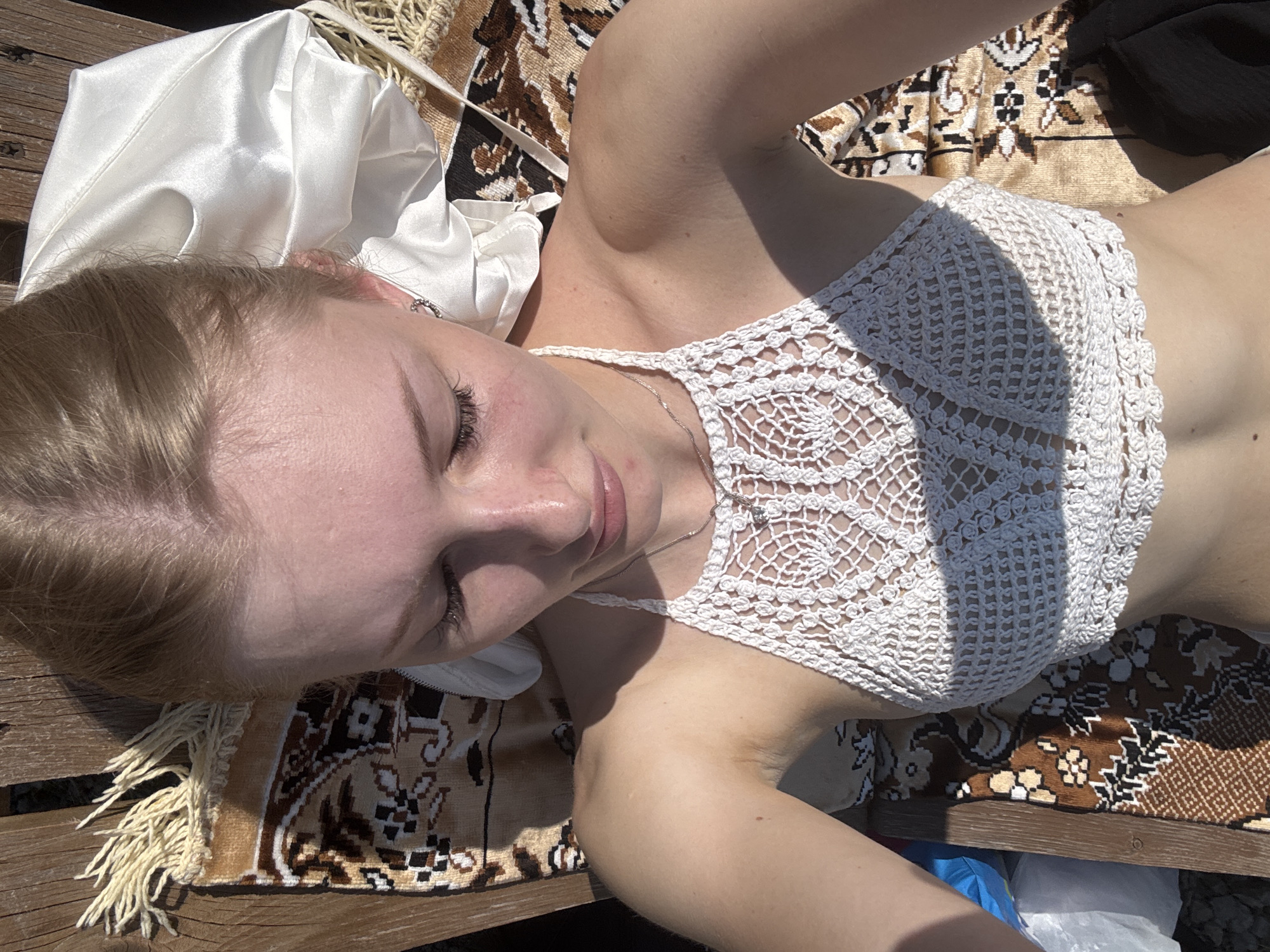 A young blonde woman with fair skin and closed eyes is lying on a wooden bench outdoors. She is wearing a white crochet halter top and a silver necklace. A patterned rug with a fringe is spread on the bench beneath her, and a white silky fabric is draped near her head. Sunlight casts shadows across the scene. The bench is situated on a surface that appears to be made of gravel. In the bottom left corner, a blue plastic object and a white plastic bag are visible. The environment suggests an outdoor setting, possibly a park or a backyard in the city of Karyer Izvestnyak, Russia.