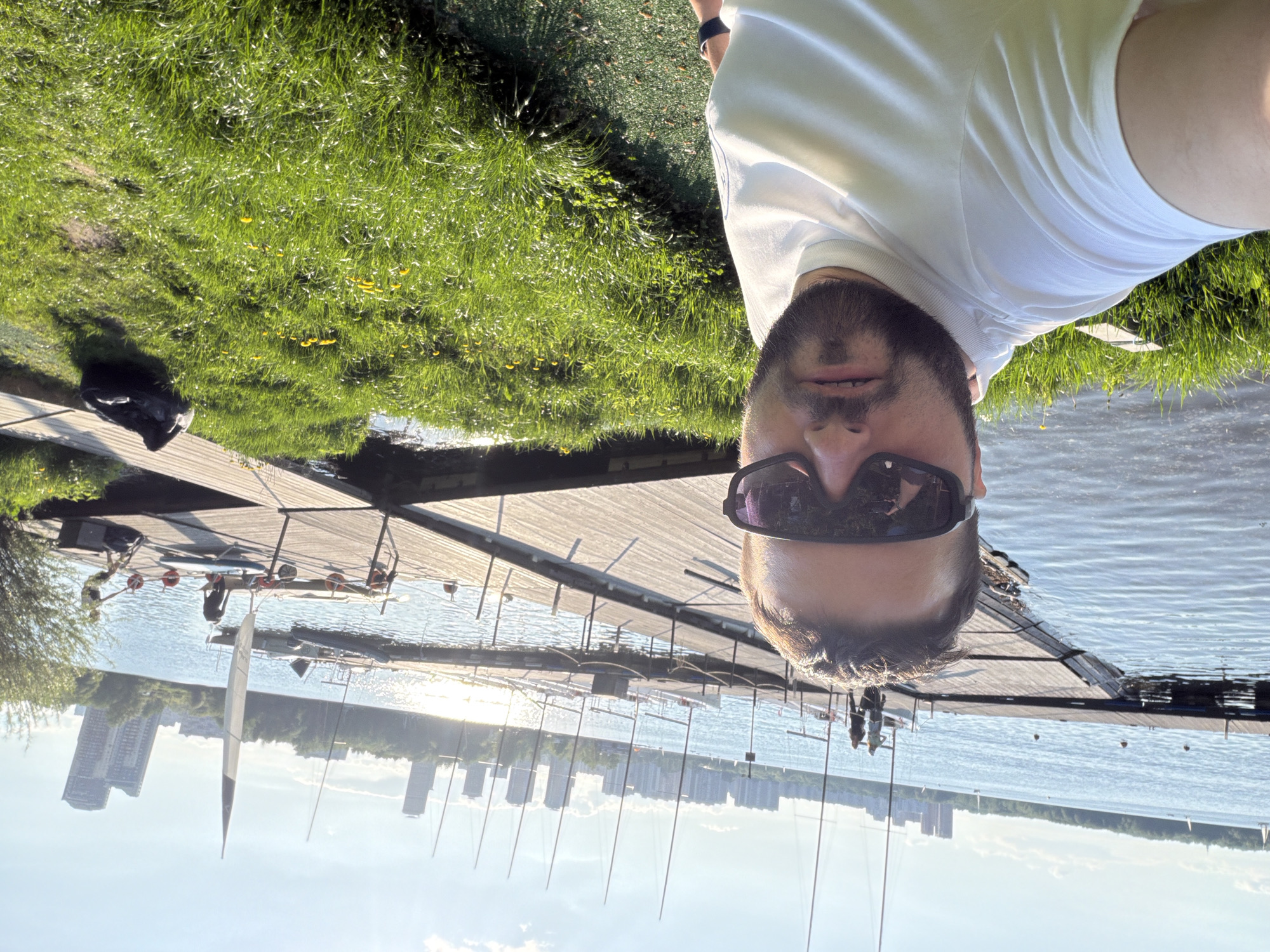 A selfie of a man with a beard and sunglasses, wearing a white shirt, taken outdoors on a sunny day in Khimki, Russia. The man is standing on a wooden dock or pier, with green grass and water visible. In the background, a cityscape with buildings and sailboats is visible. The image is oriented so the subject is in the lower right.