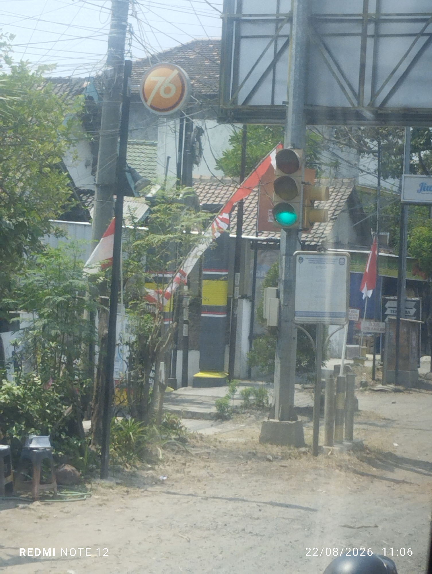 The image is a slightly blurry, medium-angle shot taken outdoors during the daytime. It appears to be a street scene in Jiwan, Indonesia, as indicated by the timestamp "22/08/2026 11:06" and the text "REDMI NOTE 12" in the bottom left corner, suggesting it was taken with a Redmi Note 12 phone.
In the foreground, a dirt and gravel path leads towards the background. To the left, there are two wooden stools. The middle ground is dominated by a traffic light pole with a green light illuminated. Several Indonesian flags, red and white, are visible, some attached to poles and others draped. Behind the flags, there are buildings with tiled roofs and some dense foliage. A circular sign with the number "76" in orange and white is mounted on a utility pole to the left of the traffic light. In the background, more buildings and trees are visible under a bright sky. The overall impression is a somewhat chaotic urban or semi-urban environment. No people are clearly visible in the frame.