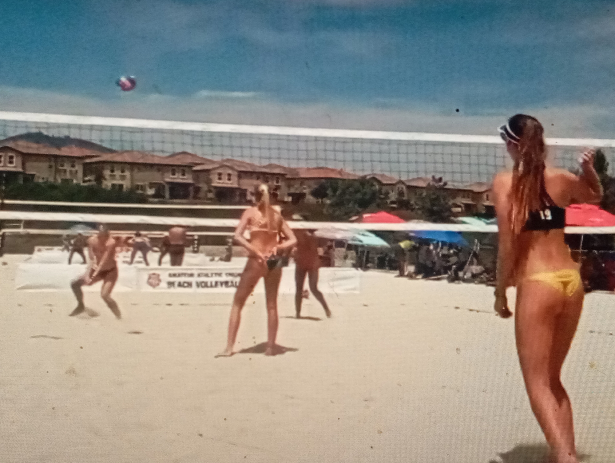 This media file depicts a beach volleyball game on a sunny day. The scene is set on a sandy beach with a volleyball net in the foreground. Several individuals are actively participating in the game, with some players positioned on either side of the net. A volleyball is visible in the air above the net, indicating that a rally is in progress. In the background, there are buildings that appear to be residences, and tents or umbrellas providing shade. The weather seems to be clear and bright, suggesting it is daytime. The atmosphere appears to be one of sportsmanship and recreation. There is text visible on a banner behind the players, which reads "BEACH VOLLEYBALL". The location is identified as Sofiivska Borshchahivka, Ukraine.
