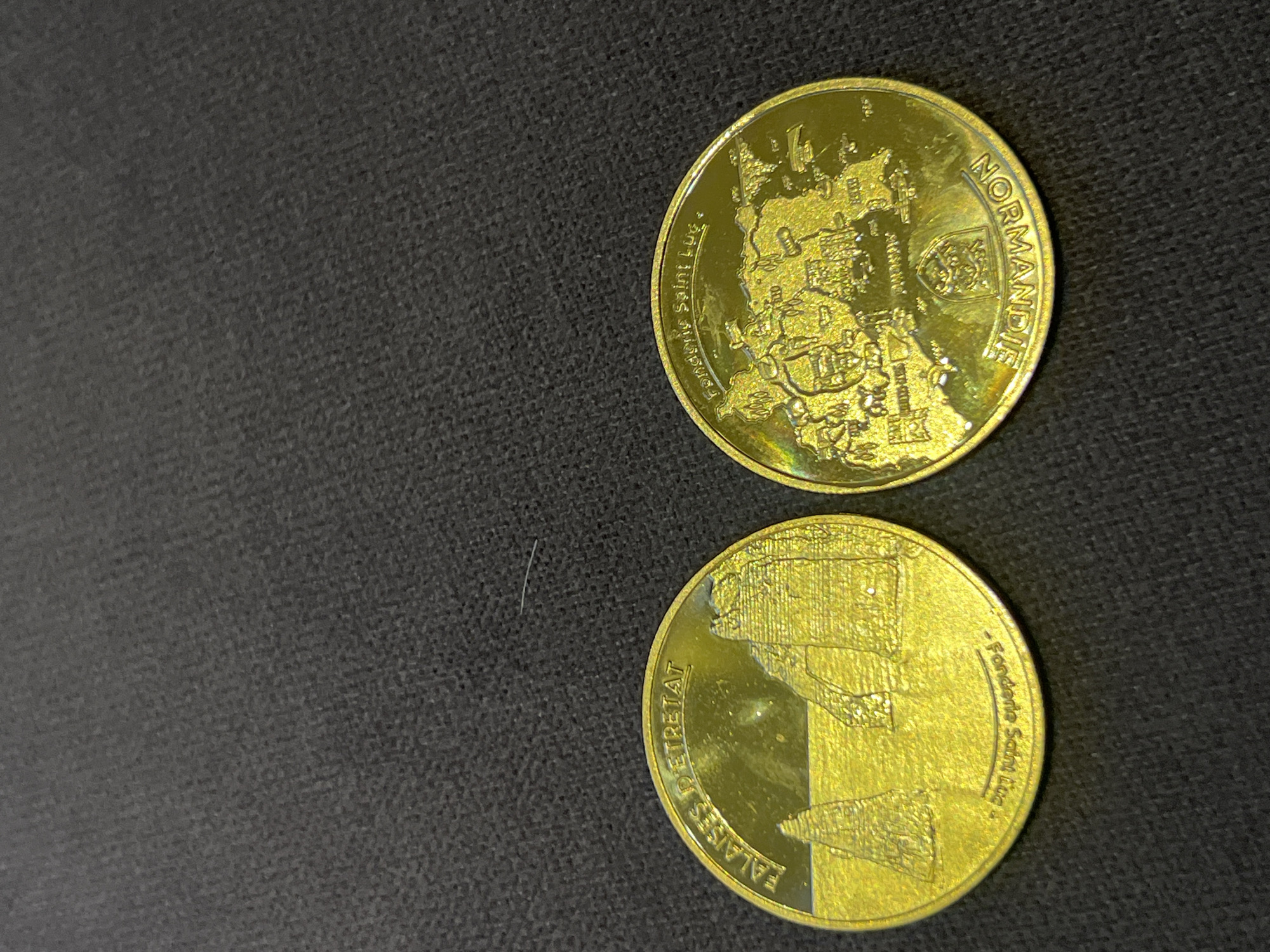 Two gold-colored commemorative coins are displayed on a dark, textured surface. The coin on the left features an engraved image of coastal cliffs with rock formations, labeled "FALAISES D'ETRETAT" along the top edge and "- Fonderie Saint Luc -" along the bottom edge. The coin on the right depicts a map-like illustration with buildings and geographical features, along with the inscription "NORMANDIE" around the bottom edge and "Fonderie Saint Luc" along the top edge. The lighting creates reflections and highlights on the metallic surfaces of the coins.