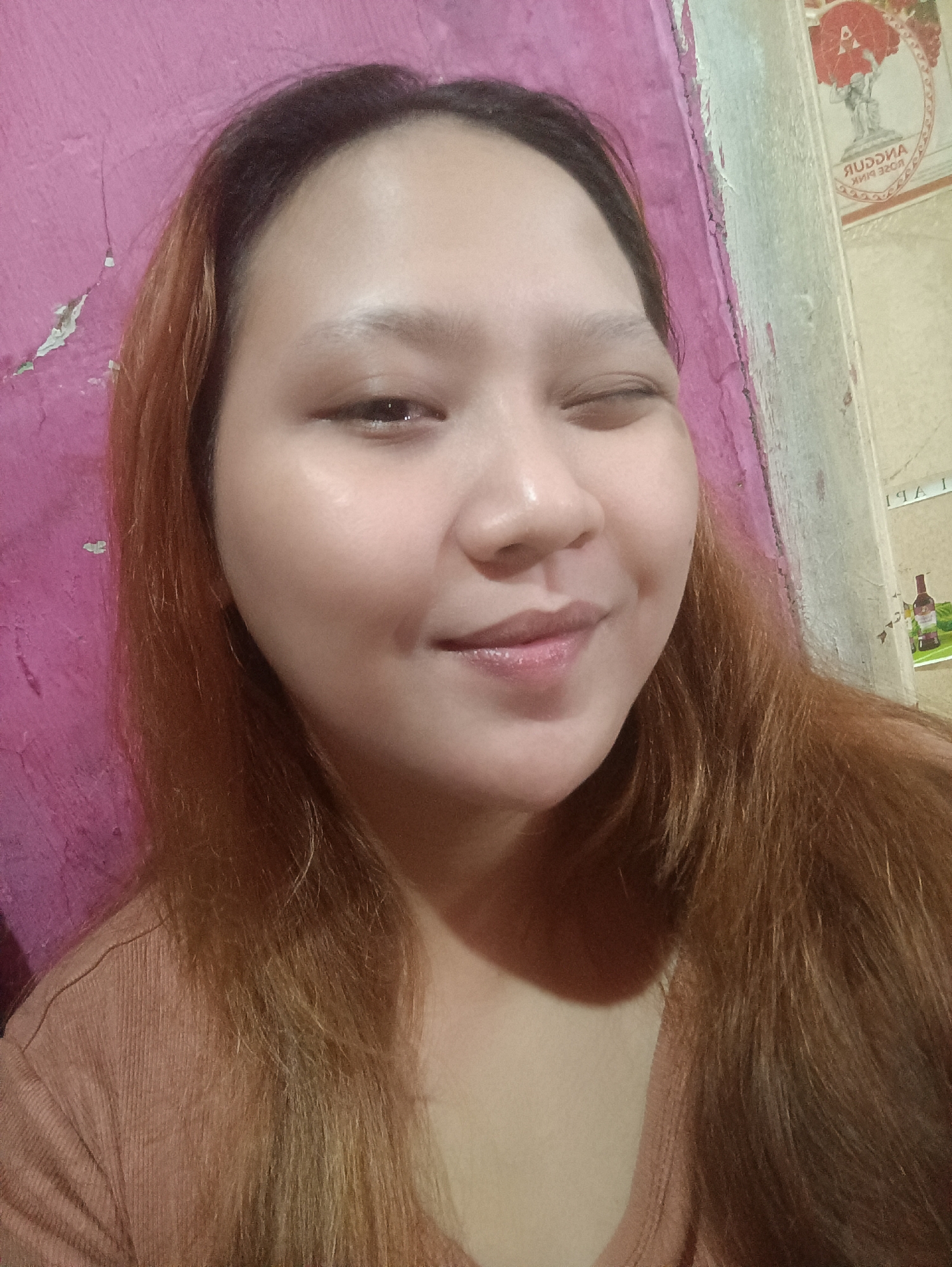 A portrait of a young woman with fair skin, auburn hair, and a pink shirt is shown. She is winking and smiling slightly. The background consists of a pink wall with some peeling paint and a lighter, textured wall with a poster that has text in Cyrillic script. The overall lighting suggests an indoor setting. The image is an unposed selfie, likely taken in a residential area of Depok, Indonesia.