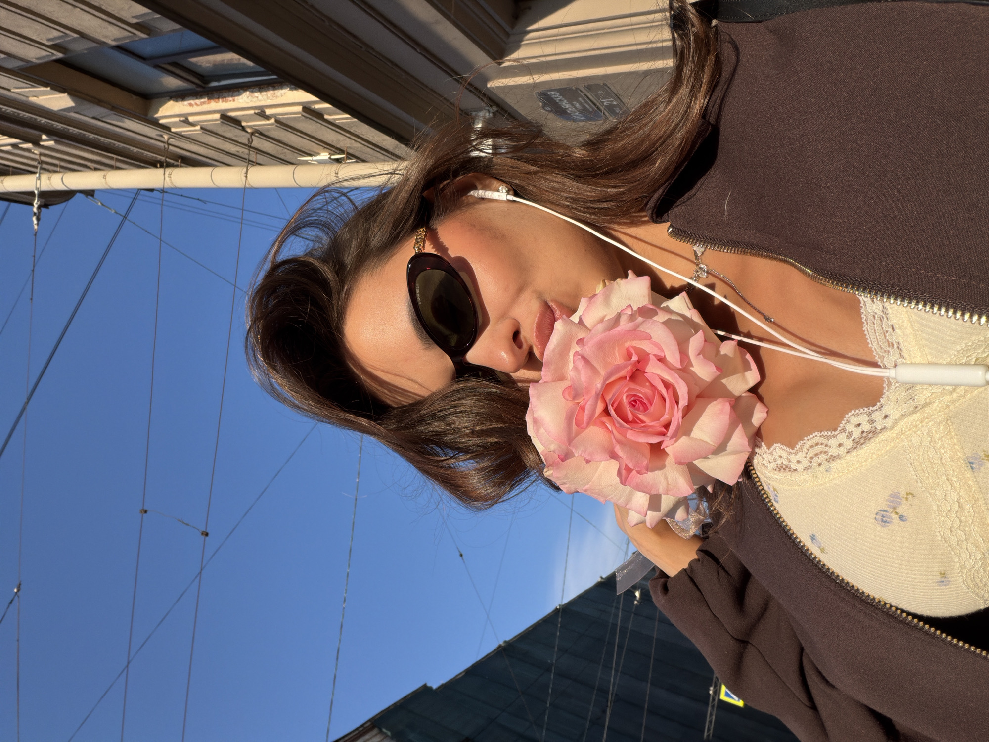 A young woman, wearing sunglasses, a brown zip-up jacket, and a cream-colored lace-trimmed top, holds a large pink rose to her face, obscuring her mouth. She is wearing white earbuds. The woman stands outdoors in what appears to be a city street, possibly in St. Petersburg, Russia, as evidenced by the architectural style of the buildings and a street sign visible in the background that reads "ИТАЛЬЯНСКАЯ 21" (Italian Street 21). The sky is clear and blue, with visible utility wires crisscrossing overhead. The sunlight casts shadows, indicating it is likely daytime. The composition is a close-up shot, focusing on the woman and the rose.